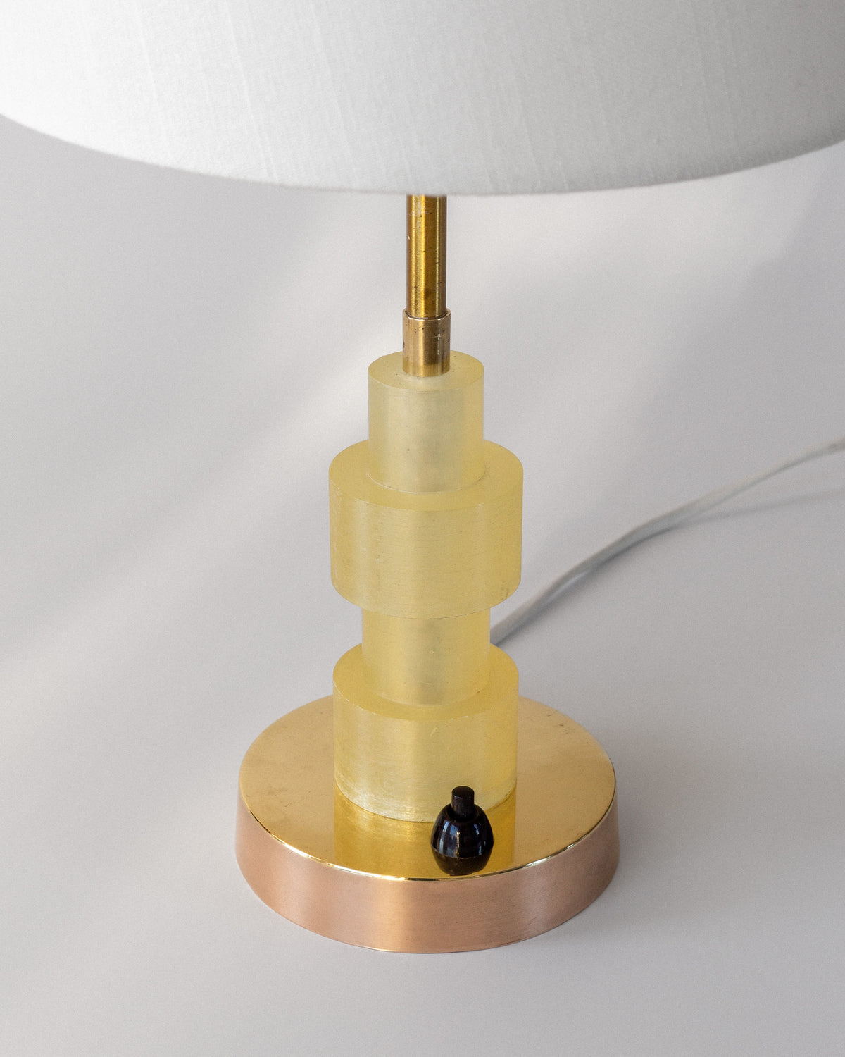 A close-up of the Spigel Swedish Mid-Century Table Lamp showcases a stacked cylindrical base in translucent yellow on a gold stand. It includes a small black switch and a partially visible white textile shade.