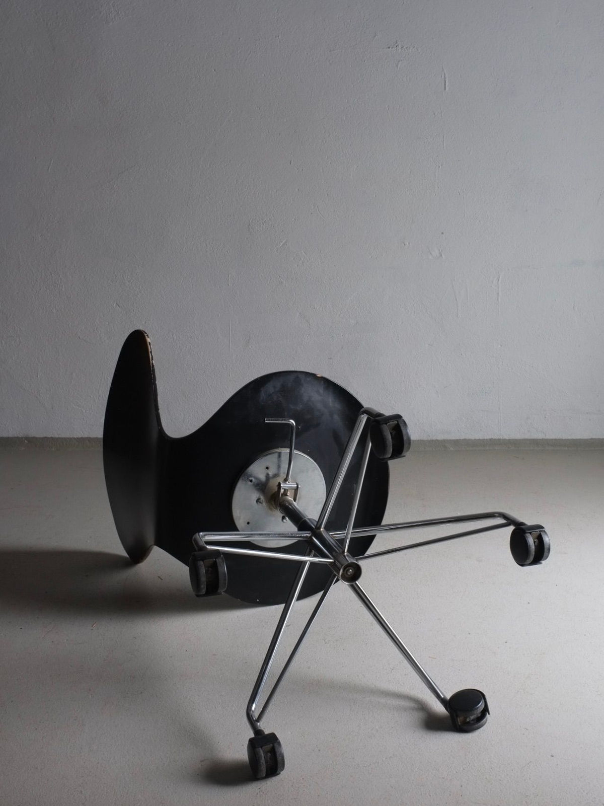 A 3117 Black Swivel Chair by Arne Jacobsen for Fritz Hansen, reminiscent of Arne Jacobsen's style and produced by Veter Vintage, lies on its side on a white floor against a plain, light gray wall. Its chrome metal base and wheels glint under the light, emphasizing that the chair has toppled over.
