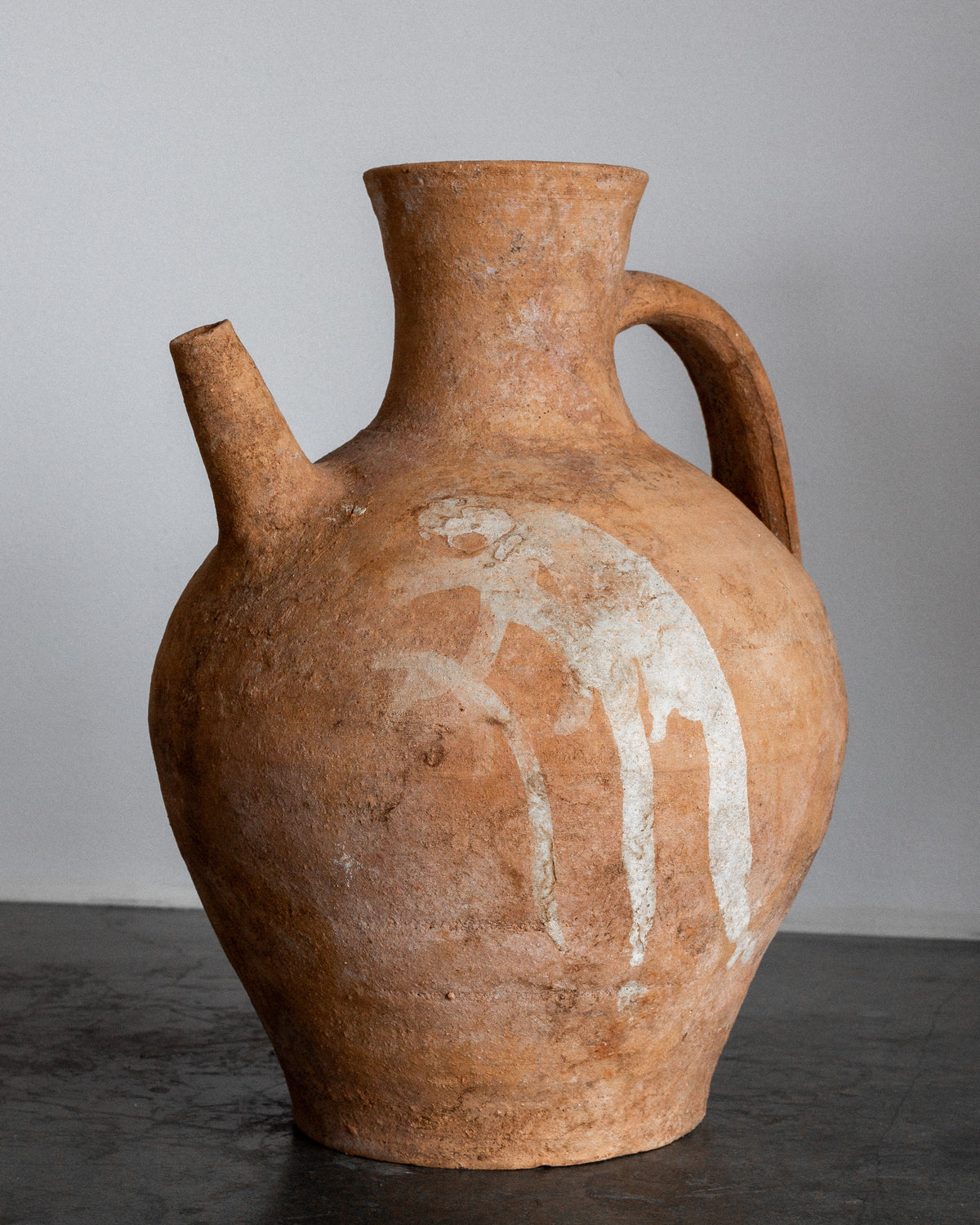 A Spigel Early 20th Century European Ceramic Pitcher, featuring a rustic hand-crafted design with a single handle and spout, stands on a gray surface. This earthenware jug has an aged texture with faint white patterns, set against a plain light gray wall.