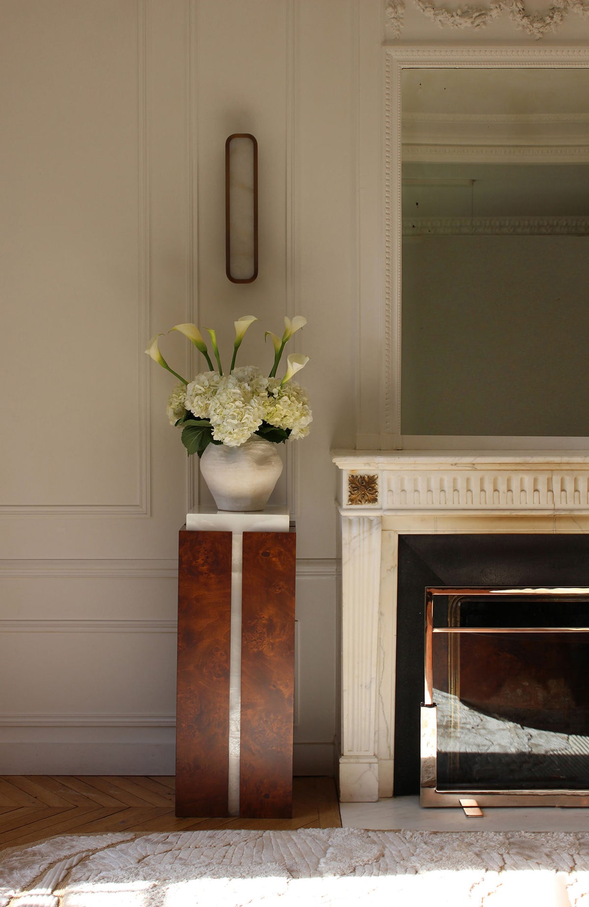 A vase of white calla lilies and hydrangeas sits atop Marbera's 'Como' Plinth in White Onyx and Brown Wood beside an ornate white fireplace with a large mirror above, brightening the elegant, sunlit room.