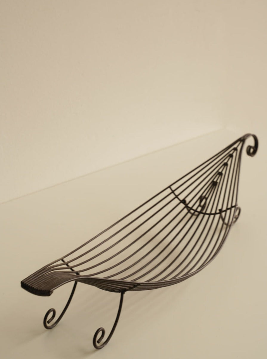 An Iron Swirl Fruit Bowl by Les Objets Amsterdam, designed to resemble a boat with curved lines and a lattice pattern, is placed on a light-colored surface. The elegant fruit bowl has raised ends and sits on small spiral-shaped legs, giving it added charm with its swirl accents.