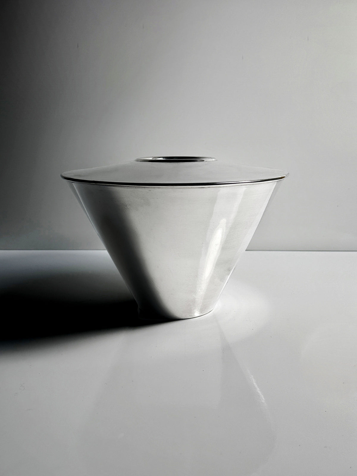 The Heavy Silver-Plated Vase from Dodo Vintage by Fink Germany, conical in design, sits elegantly on a reflective white surface. Its 1980s-inspired allure is highlighted by the lighting which casts soft shadows.