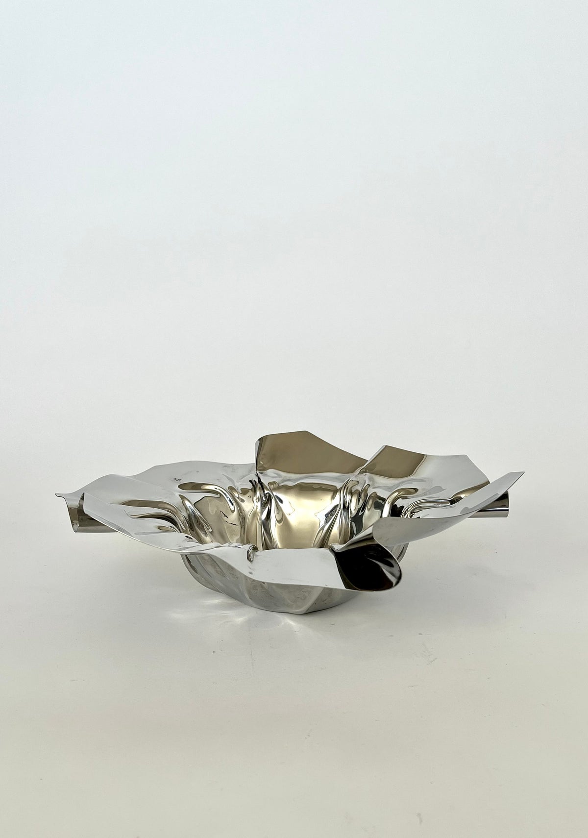 The Folded Chrome Bowl by septembre studios features a modern, reflective silver finish and an abstract, folded shape, resting elegantly on a plain, light-colored surface. Its artistic, asymmetrical design captures attention with an eye-catching visual effect, making it a striking addition to any interior setting.