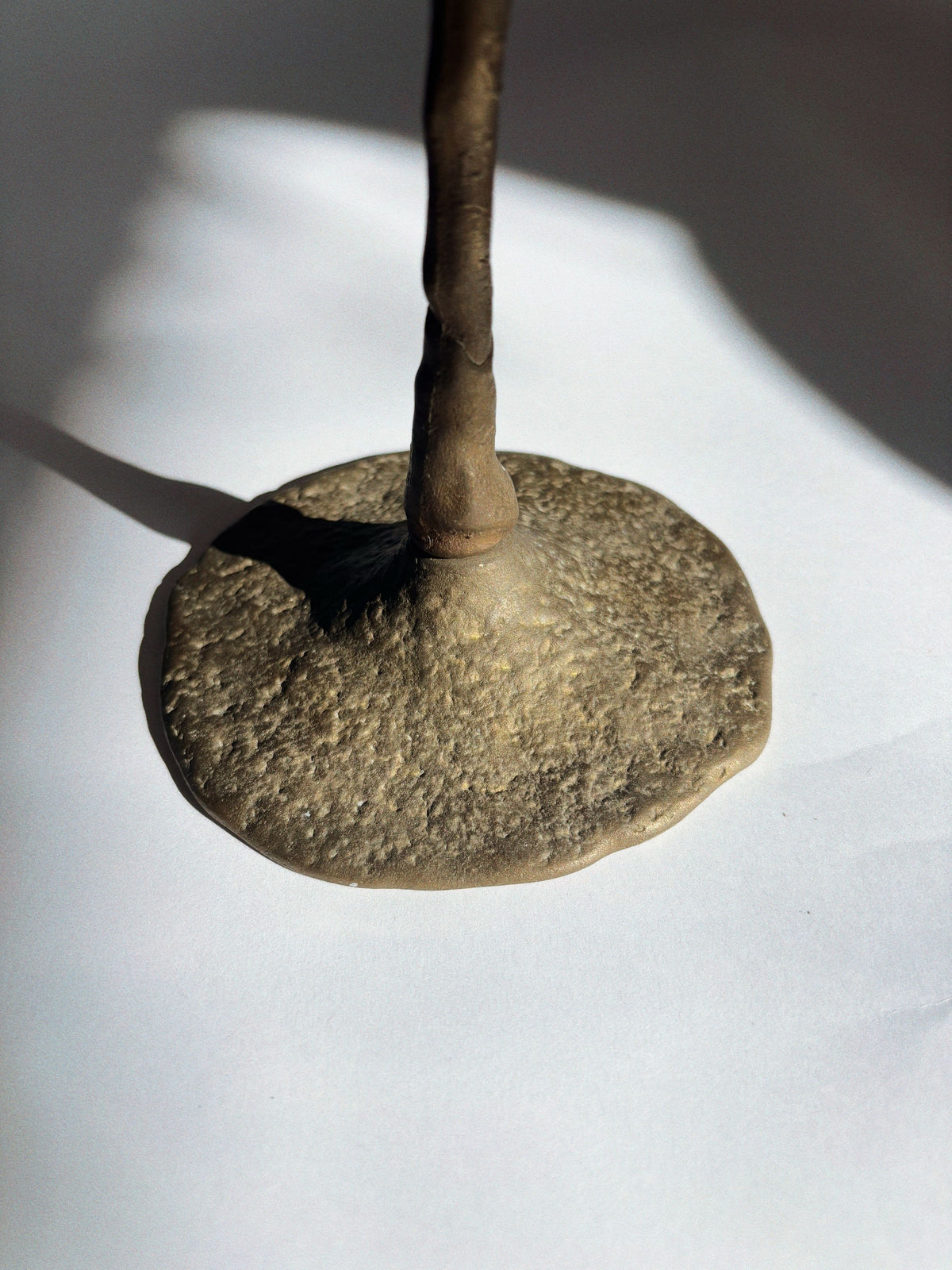 A close-up of the Huerto 2 Arm Bronze Flower Candelabra shows its slender vertical rod and textured rounded base on a white surface, casting a distinct shadow to the left.