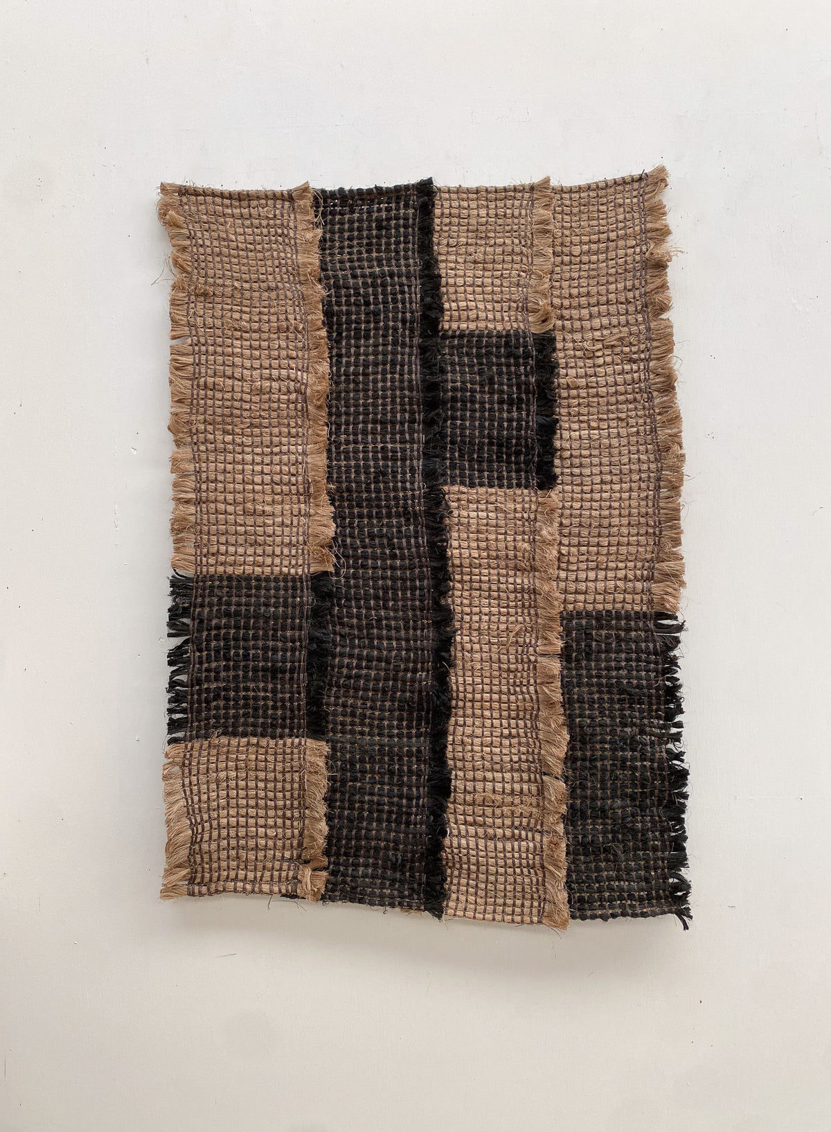 The Ink / Sand from Sundance Studio is a woven textile crafted from banana fiber, featuring a grid pattern with alternating light and dark brown sections. Its frayed edges enhance its rustic texture and highlight its biodegradable qualities.