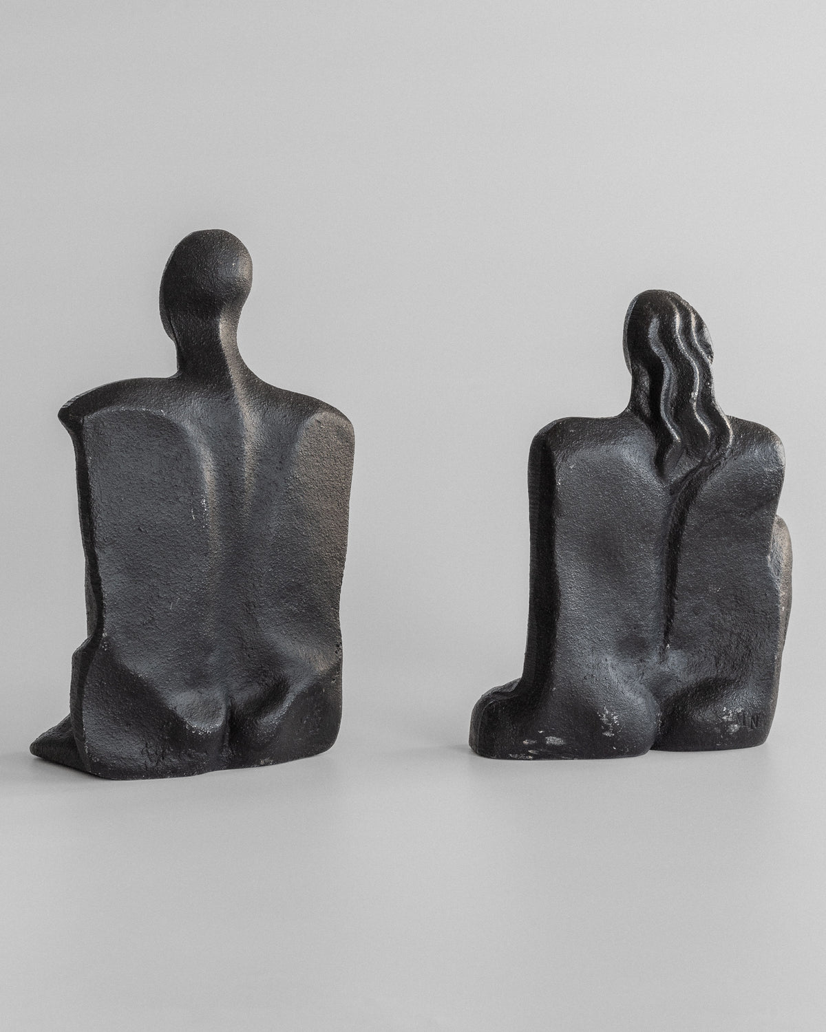 The Male and Female Sculptures by Marita Norin, from Spigel, are abstract minimalist cast-iron bookends on a gray background. The left is smooth while the right features wavy lines like hair, resembling seated human forms without detailed features—a striking artistic pair.