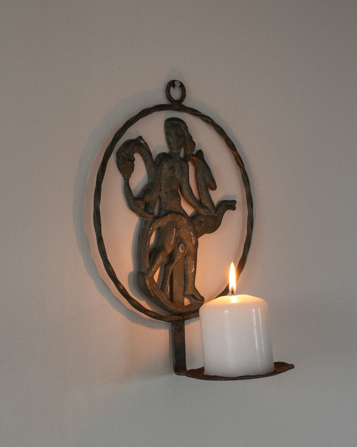 A Spigel wrought iron wall hanging has a circular design with a figure and animal, paired with an Art Deco sconce (Sweden 1920s/30s) holding a lit white candle on a plain wall, exuding vintage warmth.