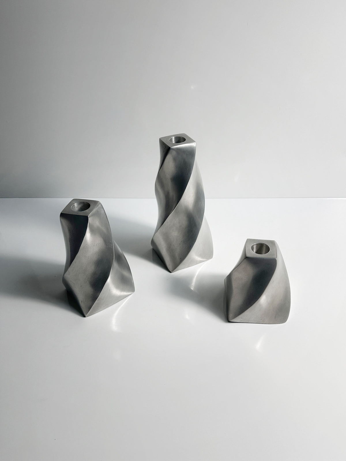 A set of three elegant Twisted Candle Holders from Dodo Vintage adorns a white surface, crafted from sleek aluminum. Each holder features unique abstract designs with smooth curves and edges in a polished finish, reflecting light and casting subtle shadows.