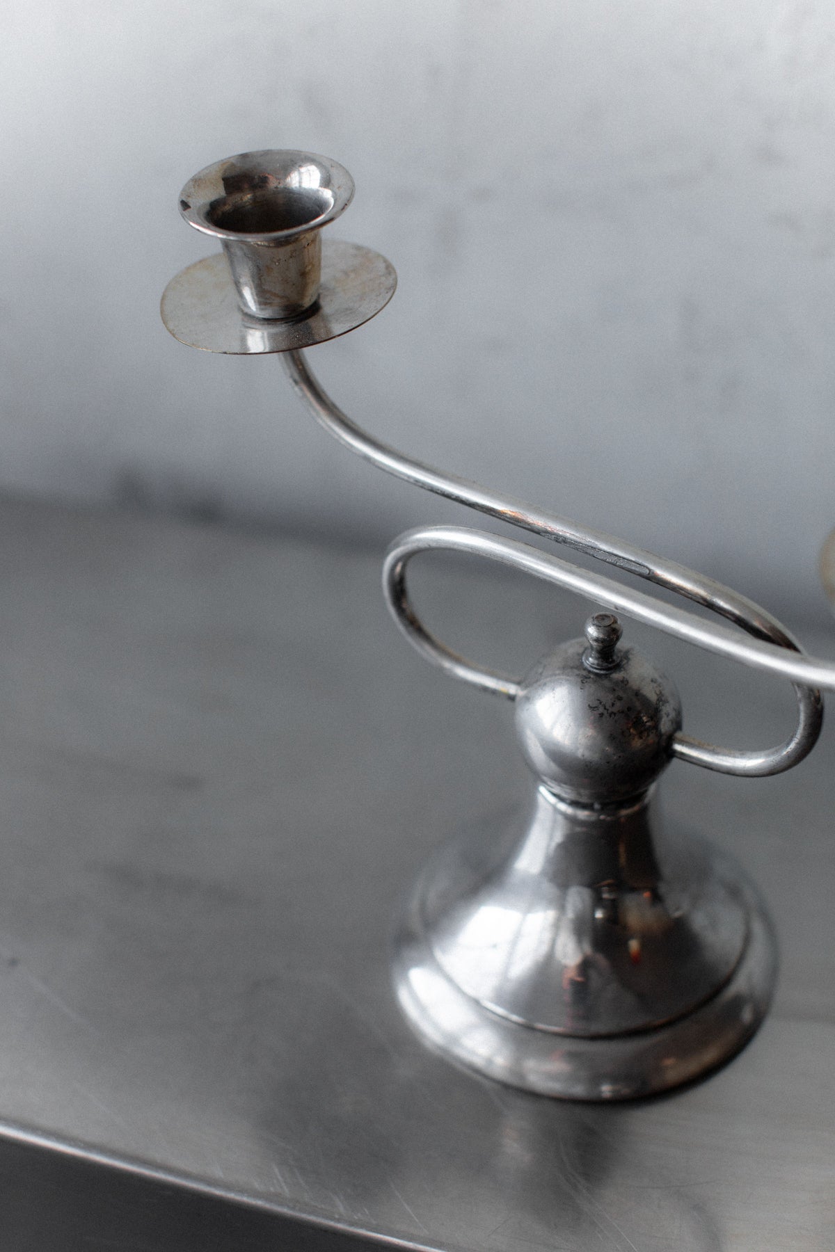 The Art Deco Candleholders by Out For Lunch exude geometric elegance with their unique, curved design as they sit on a reflective metallic surface. Their slightly tarnished finish enhances their antique charm, and the softly blurred background emphasizes these vintage-inspired pieces.