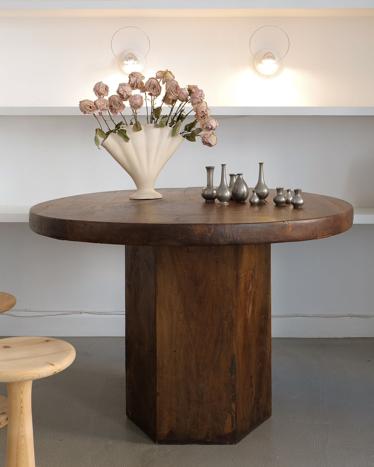 A Midcentury Hexagonal Wood Dining Table from Collection apart features a sturdy wooden base. It is adorned with a unique white vase of dried roses, and nearby, small metal pitchers enhance the minimalist white setting under soft overhead lighting.