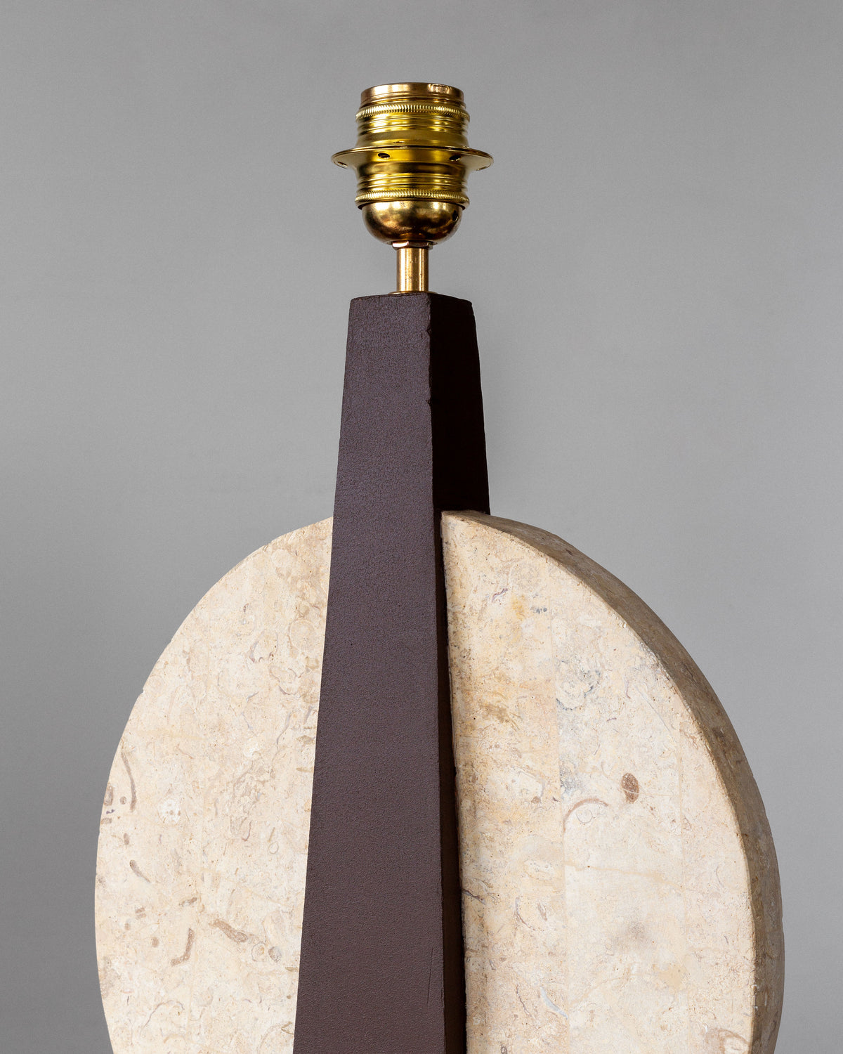The Pair of Large Travertine Table Lamps by Spigel features a minimalist lamp base with a large circular stone accent and a tall, dark brown triangular support, exuding vintage allure. It has a centrally placed brass holder reminiscent of elegant 1970s lamps against a plain light gray background.