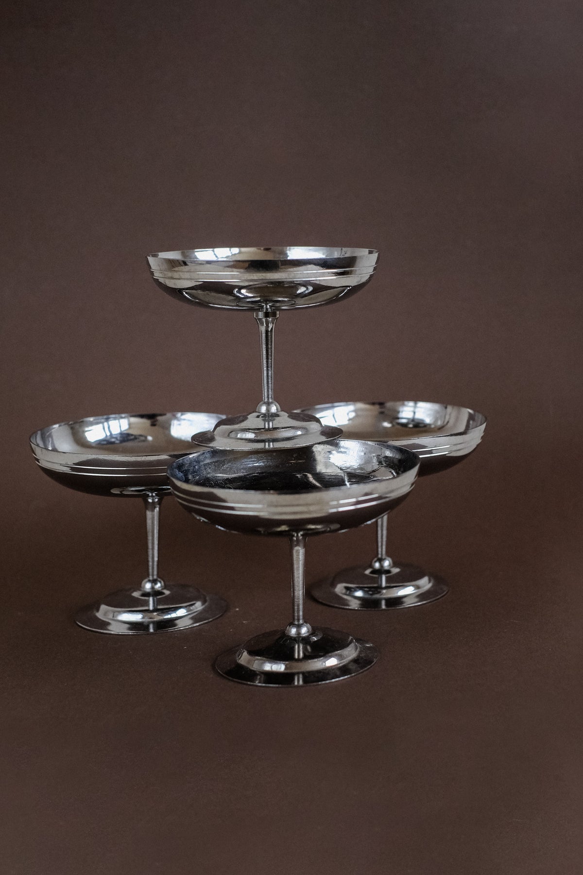 The Set of four Ice Coupes by septembre studios are French-made silver glasses with wide bowls and slender stems, beautifully reflecting light on a brown surface. They feature a classic, elegant design ideal for serving chilled drinks or ice cream.