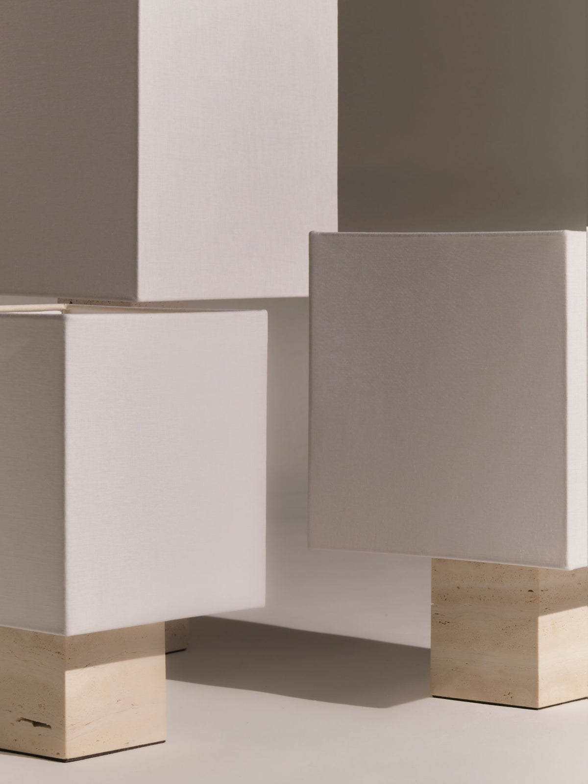 Minimalist white cube-like structures with light beige bases, placed in an abstract arrangement. The cubes vary in size and are displayed against a neutral background, showcasing geometric and clean lines. The modern composition evokes the purity of Nassi Lamps' Roma I Squared White Travertine and the elegance of white travertine.