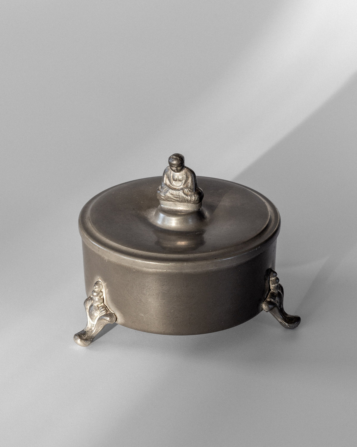 The Spigel Art Deco Metal Box from 1934 is a round pewter container with a lid adorned by a small Buddha figurine. It evokes Svenskt Tenn design aesthetics, standing on three intricately designed legs that cast gentle shadows on a light gray background.