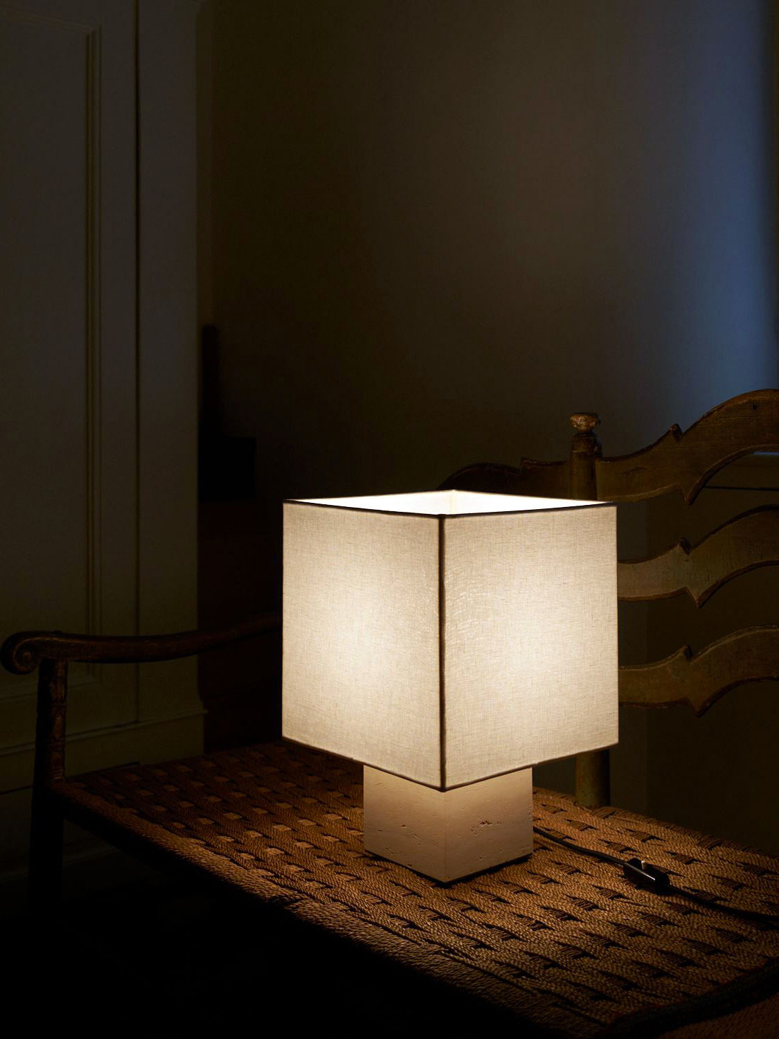 A Nassi Lamps Roma I Squared White Travertine emits a warm light, resting on a woven surface. Behind it, a wooden chair with curved armrests is partially visible in a dimly lit room. The ambiance is cozy and inviting, enhanced by elements of modernism throughout the space.