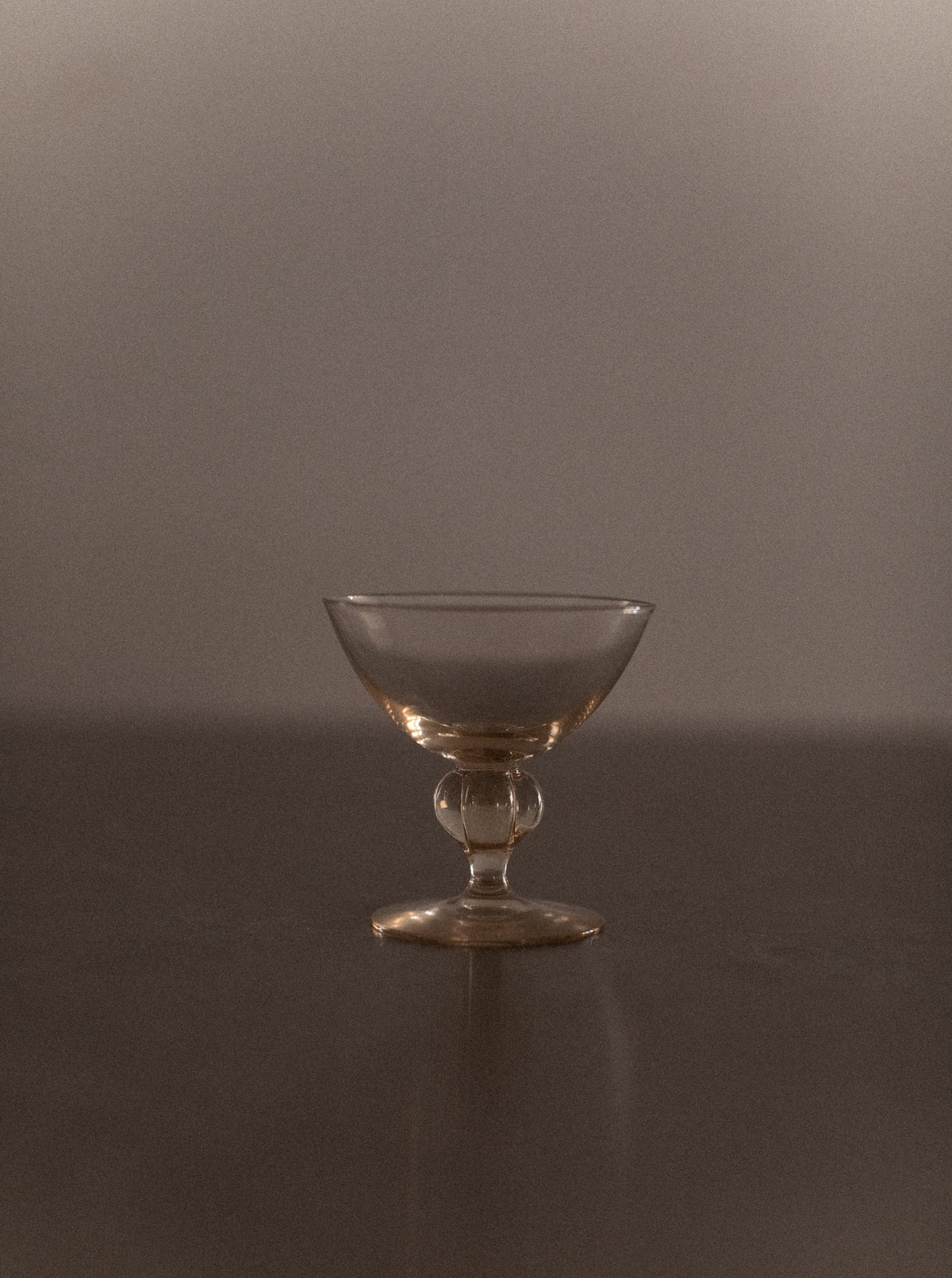 An elegant, empty goblet from the Set of 7 Art Deco Glass by Out For Lunch with a short stem sits on a flat, dark surface. The soft lighting casts gentle shadows, giving the scene a sophisticated, vintage feel. The neutral and slightly blurry background focuses attention on the glass, enhancing the anticipated drinking experience.