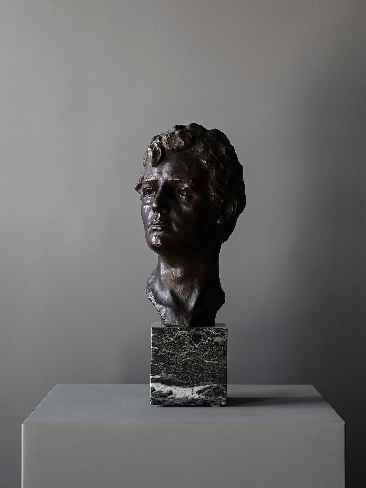The "Bronze Bust of Male by Victor Demanet" from Studio OSKLO features a man's curly-haired head on a square marble base, displayed atop a gray pedestal against a plain gray background.