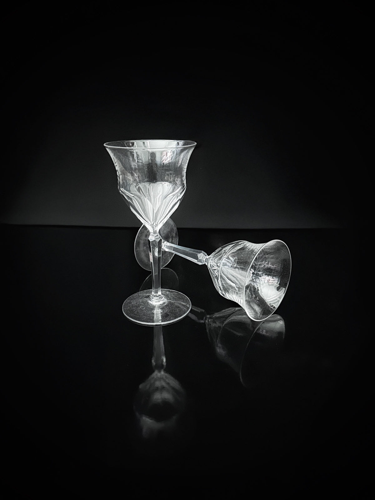 Two exquisite 9 hand cut crystal wine glasses from the 1960s by Dodo Vintage are artistically displayed on a reflective black surface. One stands upright while the other lies on its side, set against a dark background that accentuates their remarkable craftsmanship.
