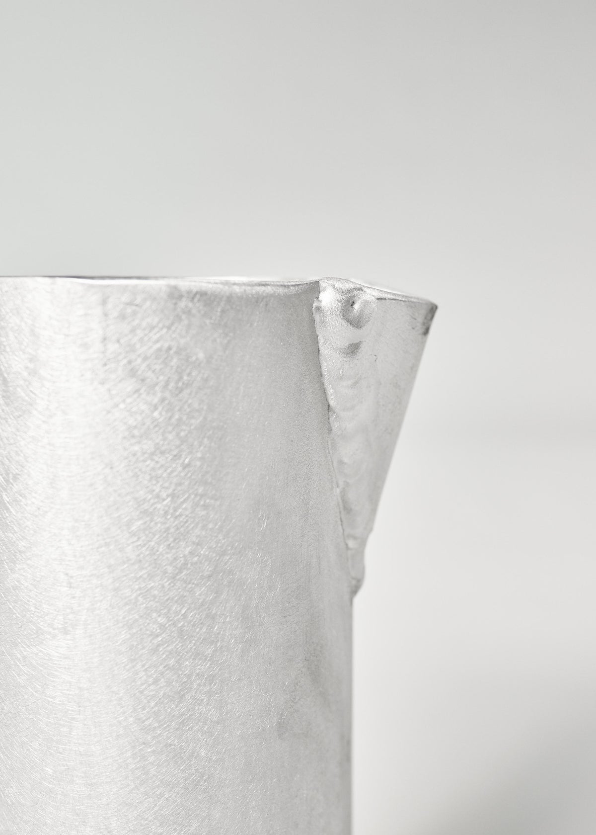 Close-up of a meticulously crafted aluminum object featuring a sleek finish and a triangular protrusion, branded as The Aluminum Jug by Six Dots Design. It is set against a soft gray background that enhances its minimalist aesthetic, akin to creations from a North London studio.