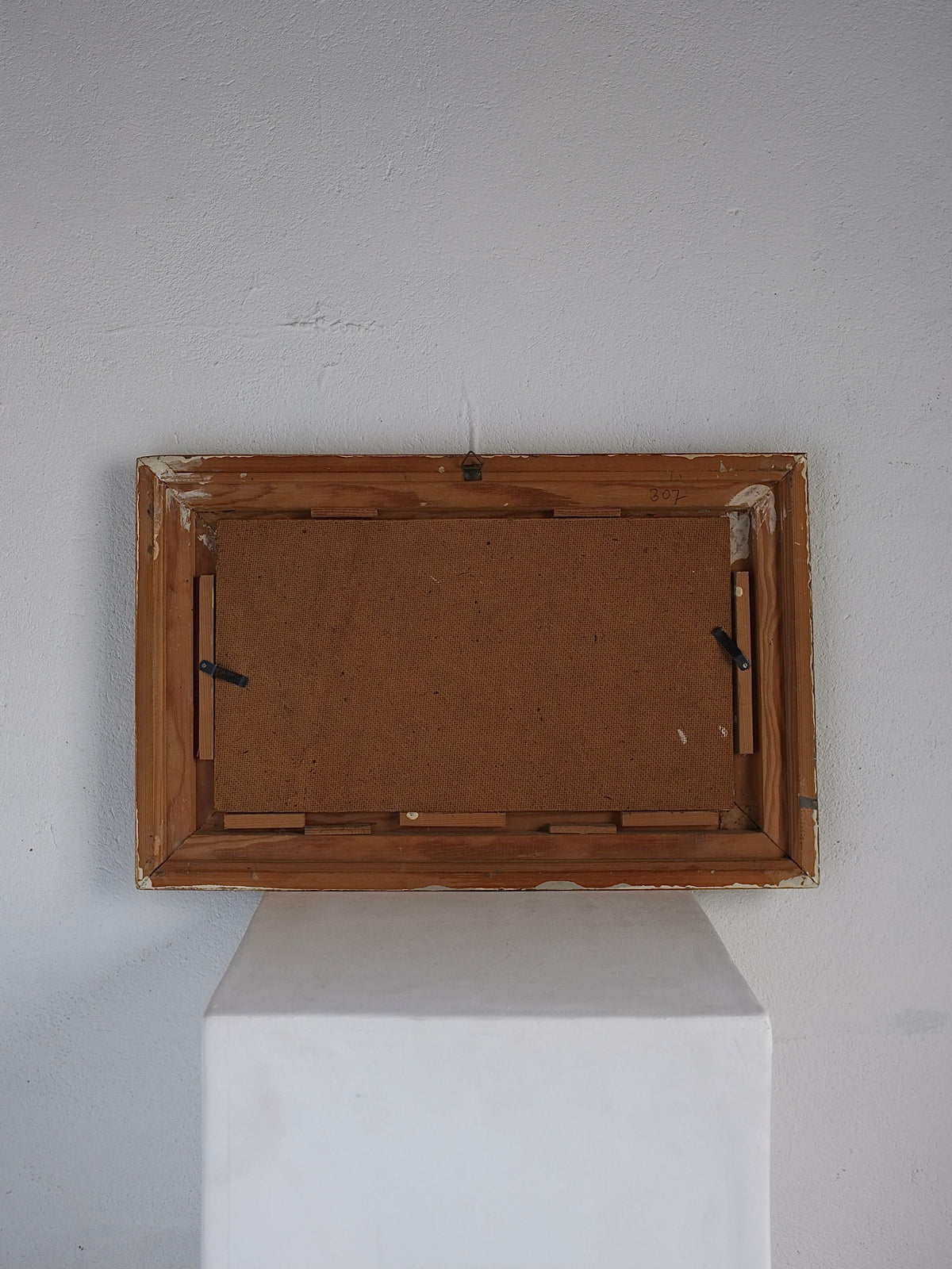 The image showcases the back of a framed artwork, likely of European origin, resting on a white pedestal against a blank white wall. The wooden frame, which seems to be in vintage condition, has a visible hook for hanging and the backing board is secured with clips. The overall setting is minimalistic and neutral. This artwork is identified as the Vintage Realism Still Life Oil Painting | 1970s by Veter Vintage.