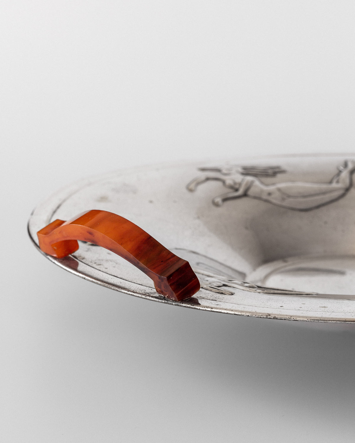 A close-up of the Decorative Bowl with Mermaids by Spigel, dating back to the 1930s, showcases an ornate design featuring mermaid engravings on its surface. The silver-plated bowl is complemented by a distinctive orange semi-transparent handle, merging practicality with timeless elegance.