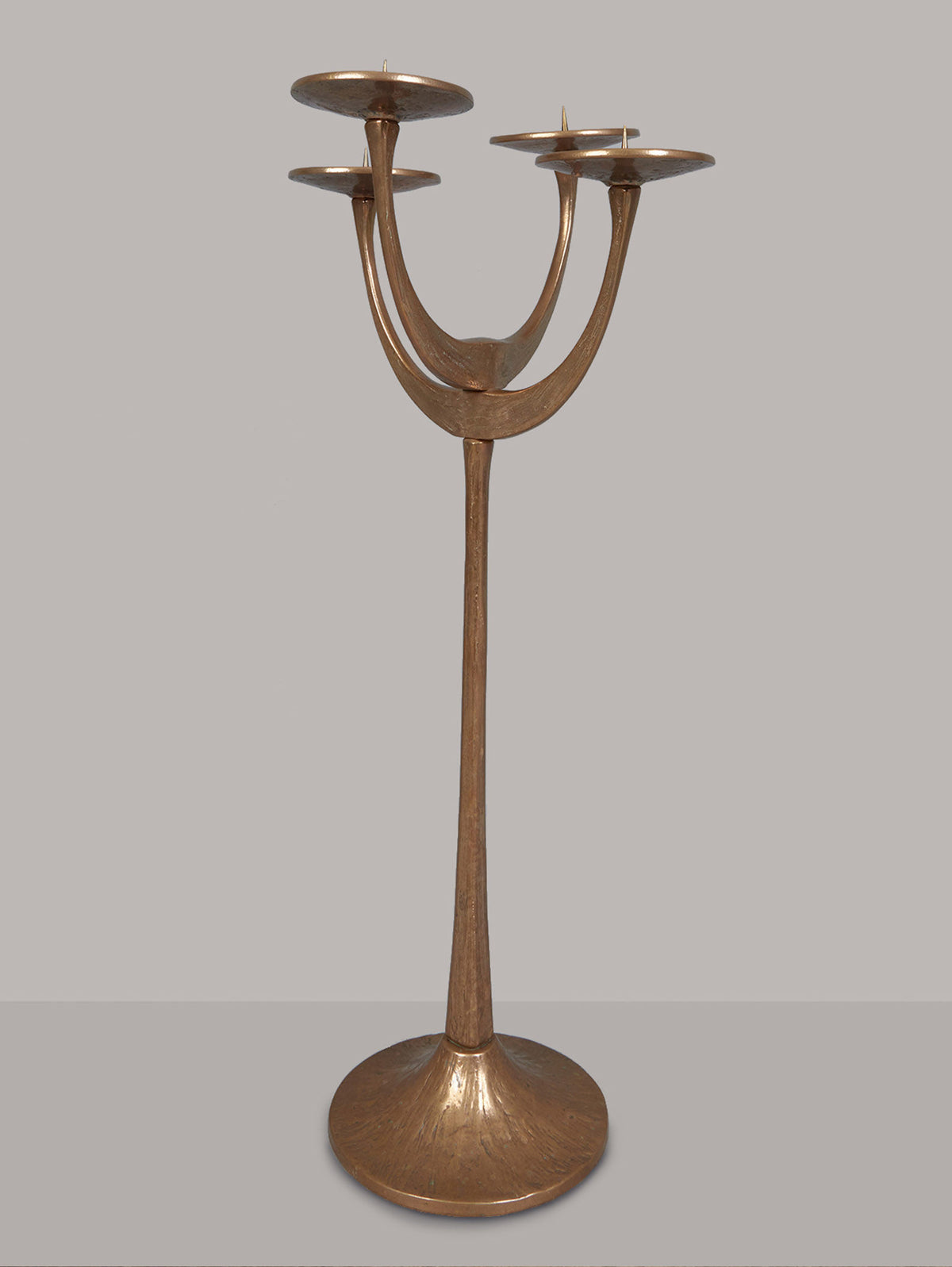 The Floor Standing Brutalist Candleholder by Tobias Harjes for RELIC LONDON features three curved arms from a central stem, each with a circular holder. Made of bell bronze, its modern design includes an elegant bronze-like finish and a wide round base for stability.