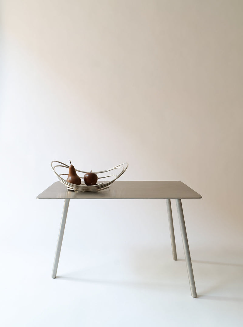 A minimalist Collection apart Stick Table Jesper Morrison 2000 with slanted legs holds a metallic fruit bowl made of Italian stainless steel, containing two pears. The background is neutral, enhancing the simple and elegant design of the table and bowl, reminiscent of a Jasper Morrison creation.