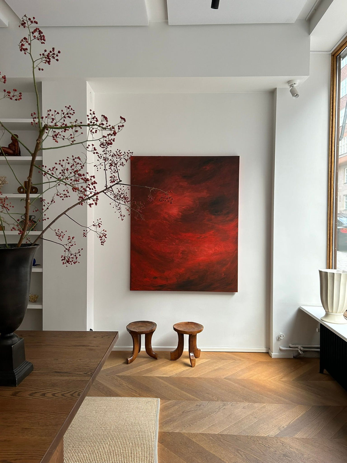 A minimalist art gallery showcases a large mixed media piece called "Ember's Whisper," designed by Mattias Storm, on a white wall. Below the artwork are two wooden stools, and to the left is a table adorned with a black vase filled with branches bearing small red berries. Natural light pours in from the window on the right.