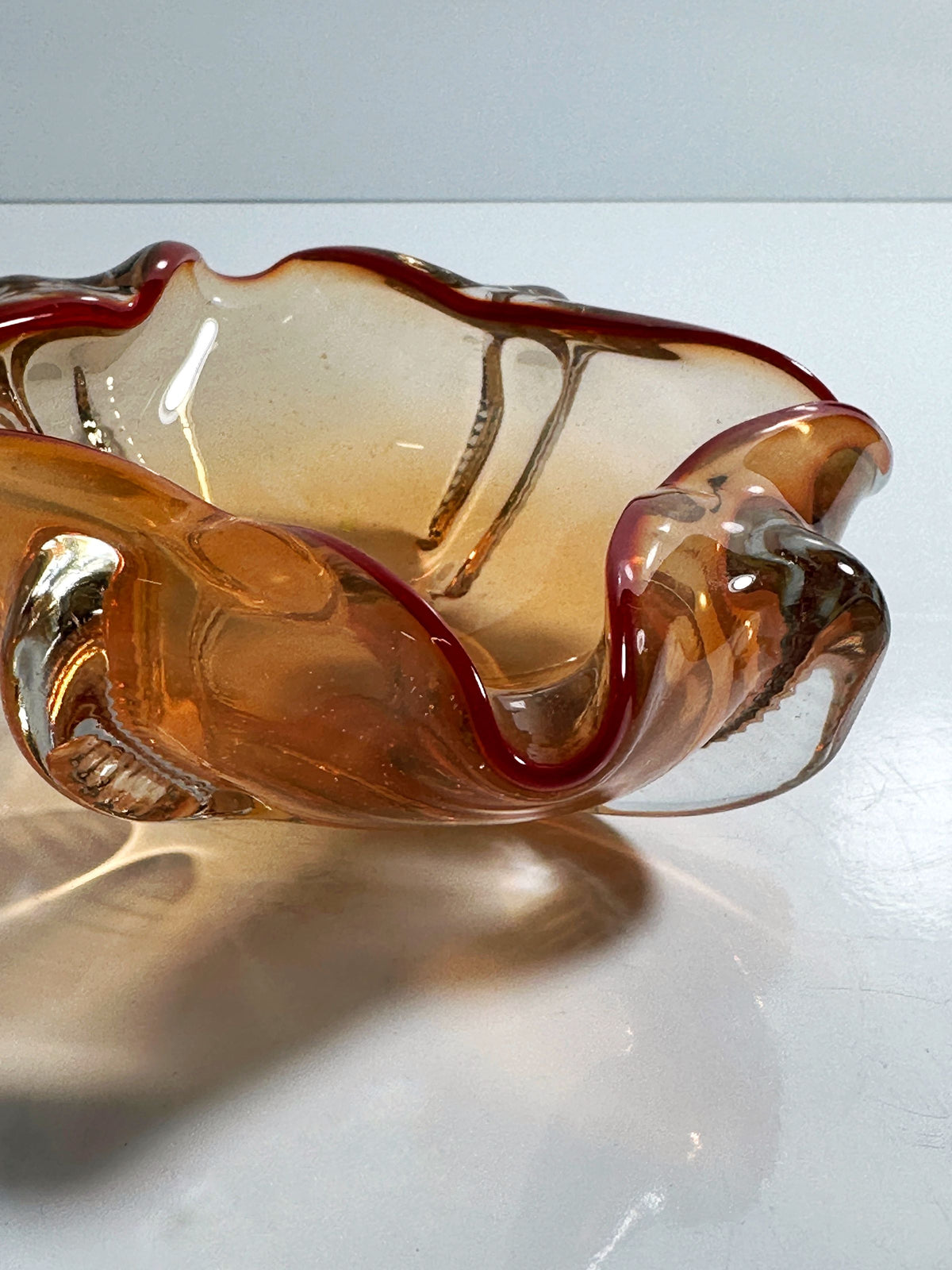 The Venetian Ashtray by Seguso Murano from the 1950s, offered by Dodo Vintage, features a wavy design and amber-clear hues. A coral-toned rim adds a red accent, while its textured surface projects delicate ripples and soft shadows on white settings, enhancing its nautical elegance.