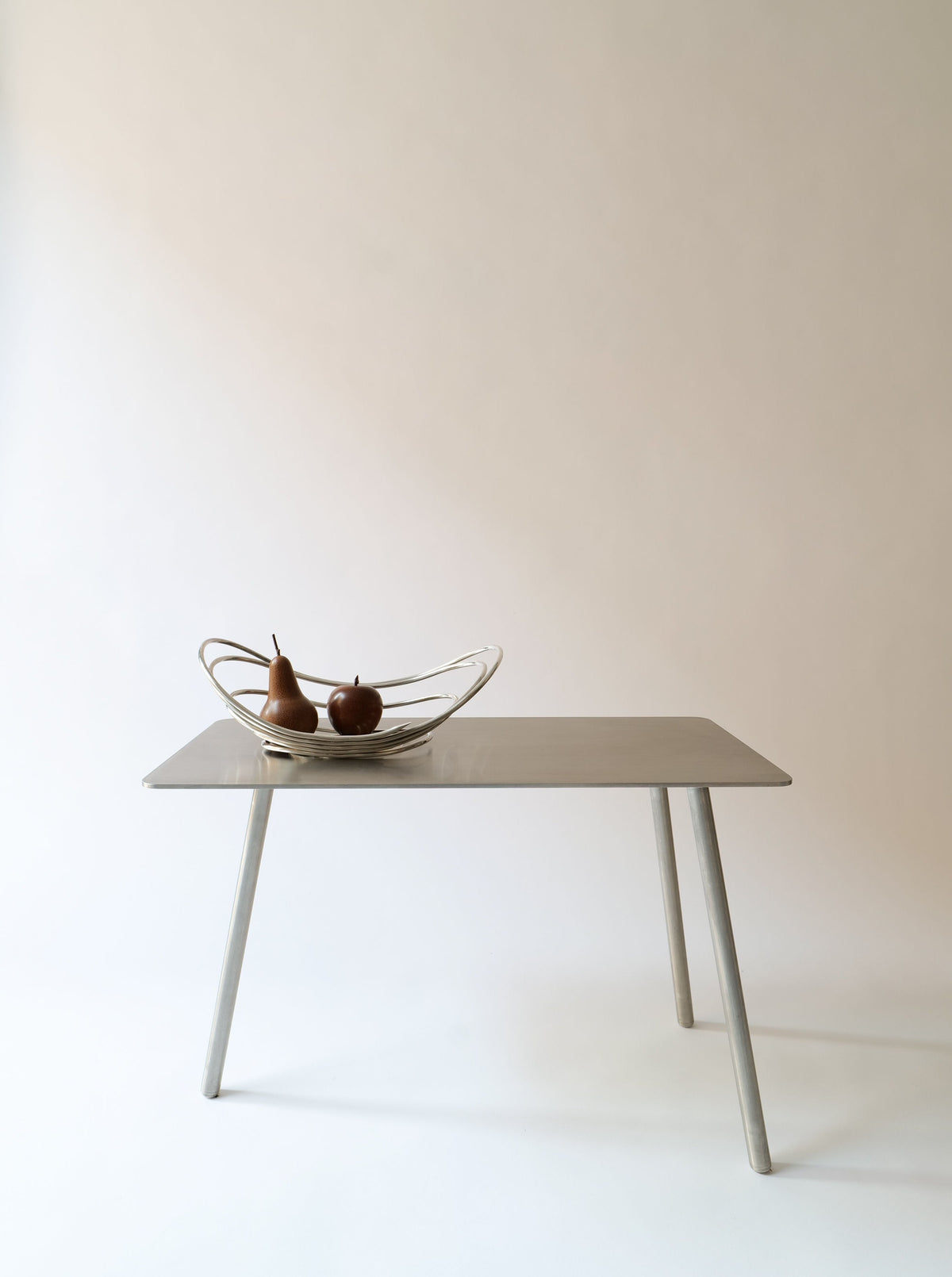 A simple, modern table with a smooth surface and four angled legs stands against a plain background. On top of the table is a decorative bowl containing two pears, serving as a Centrepiece Lino Sabattini 1970s from Collection apart. The overall aesthetic is minimalist and clean.
