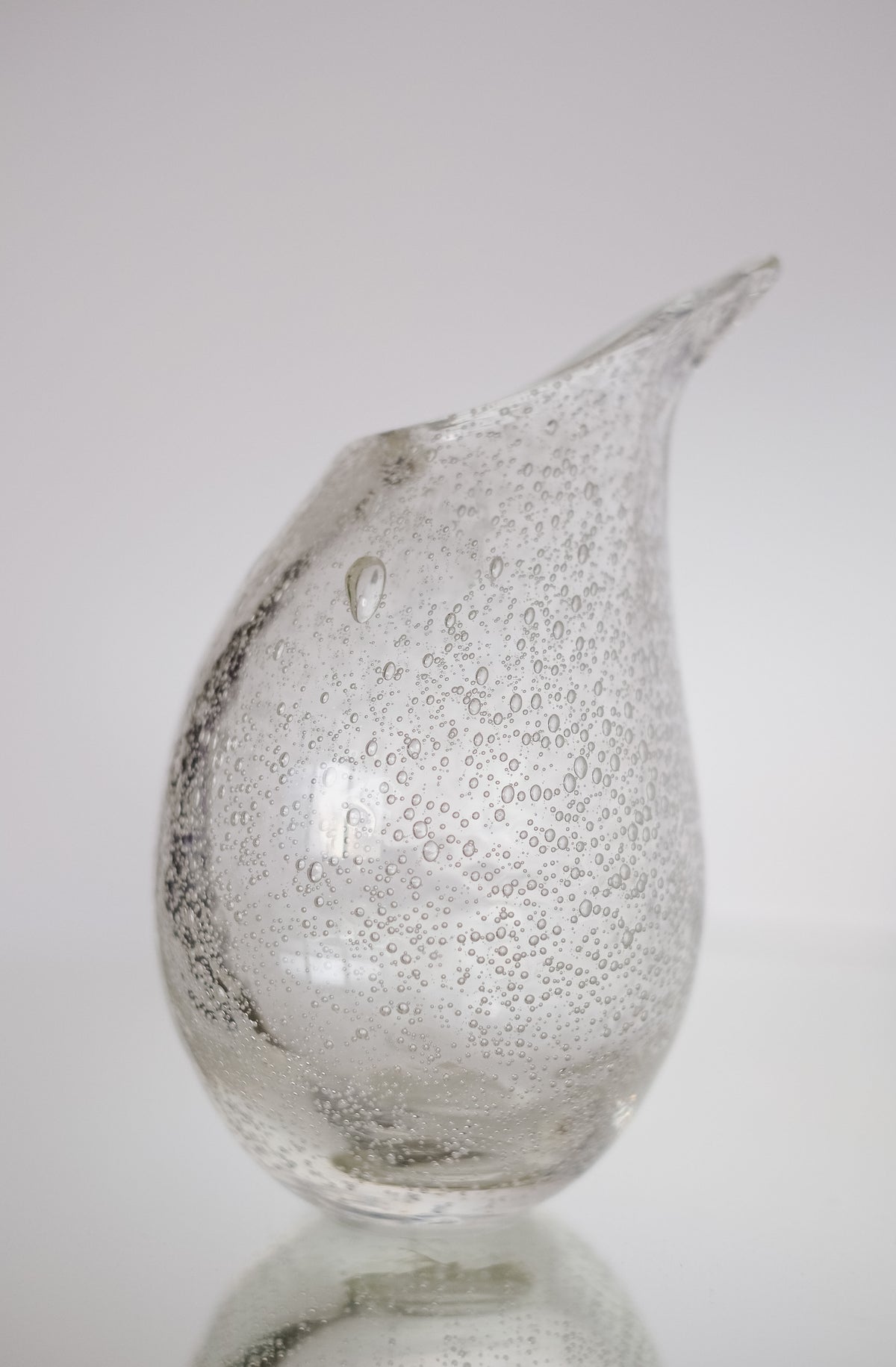 The Small Bubble Vase by septembre studios, made of transparent handmade glass with air bubbles, sits on a reflective surface against a white background. Its droplet-like form and slightly pointed tip showcase exquisite craftsmanship.
