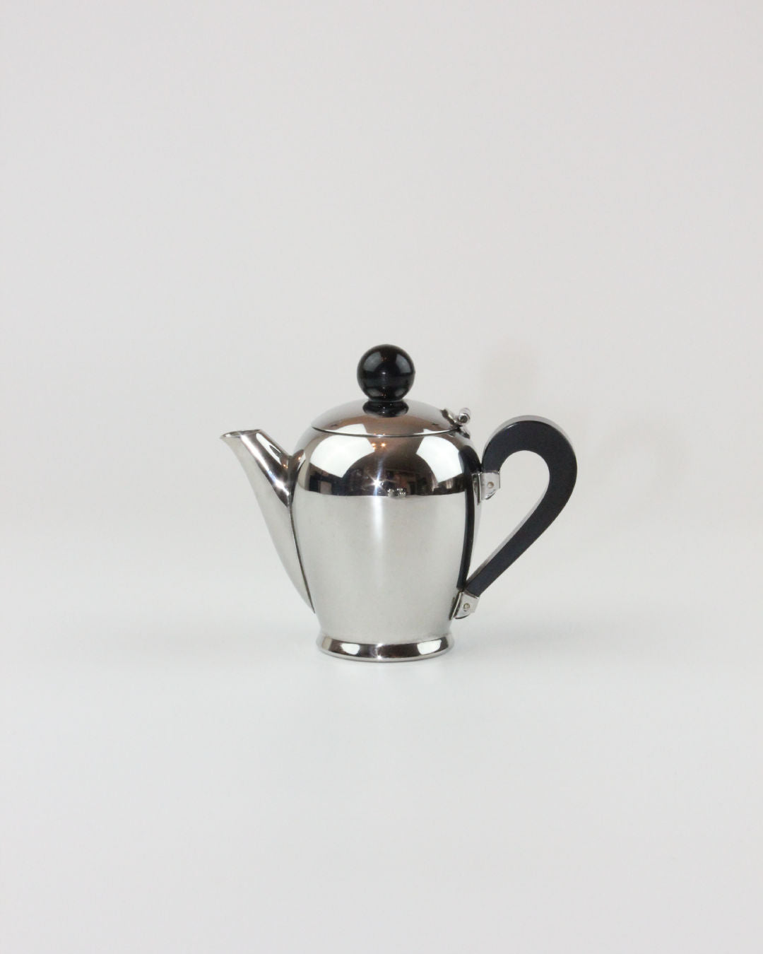 A sleek, stainless steel teapot with a glossy finish, featuring a curved spout, a black handle, and a black spherical knob on the lid, set against a plain white background. Reminiscent of mid-20th-century craftsmanship and part of the "Bombè" Alessi Tea/Coffee Set by Boga Avante Shop.