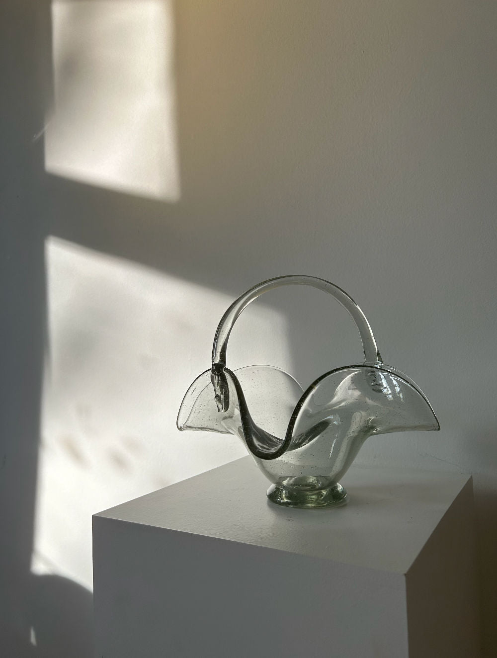 A delicate, translucent glass basket with an arching handle and leaf-like, wave-edged rims sits on a white pedestal. Soft sunlight casts a shadow on the white wall behind, creating an airy, serene atmosphere reminiscent of a 70's vintage bonbonnière. This is the **70's Vintage Glass Basket** by **Les Objets Amsterdam**.