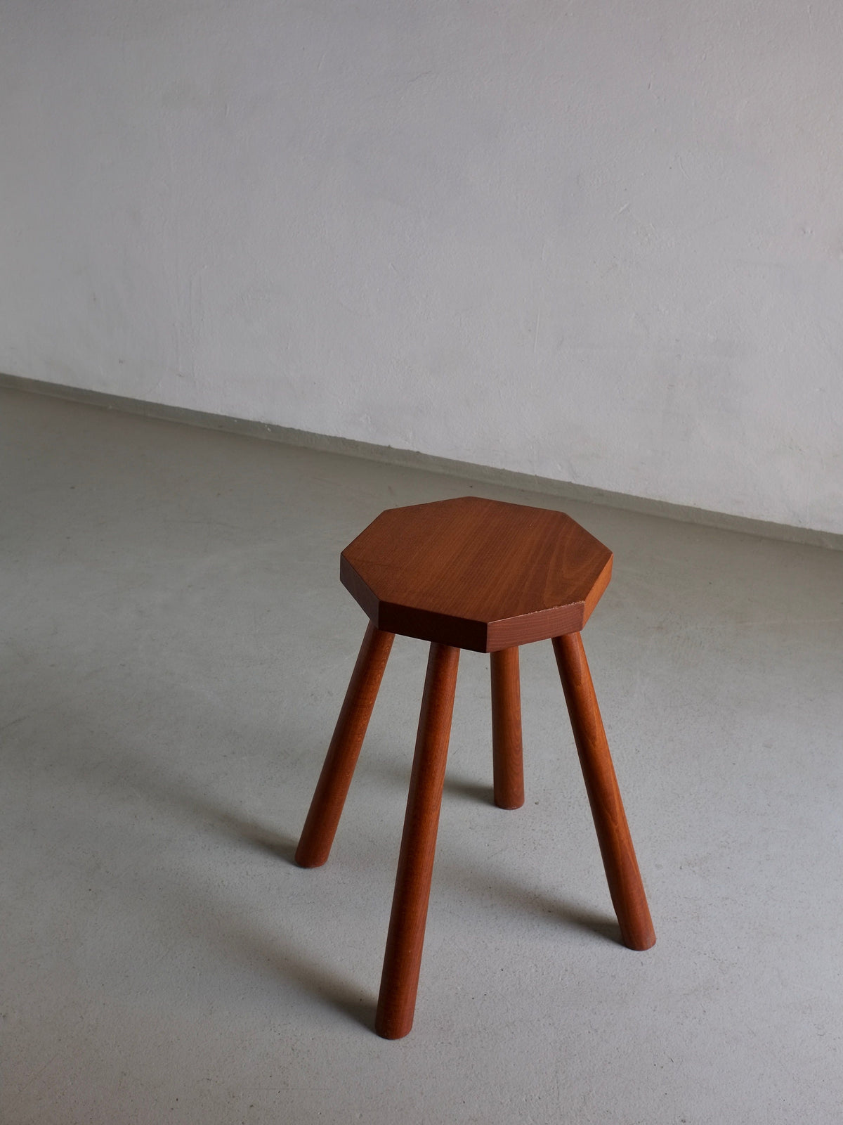 A small, vintage Solid Wood Octagonal Stool by Veter Vintage from the 1960s stands on a smooth, gray floor against a plain, light-colored wall. This minimalist side table boasts a warm brown finish with an octagonal seat and four cylindrical legs, seamlessly blending functionality with timeless style.