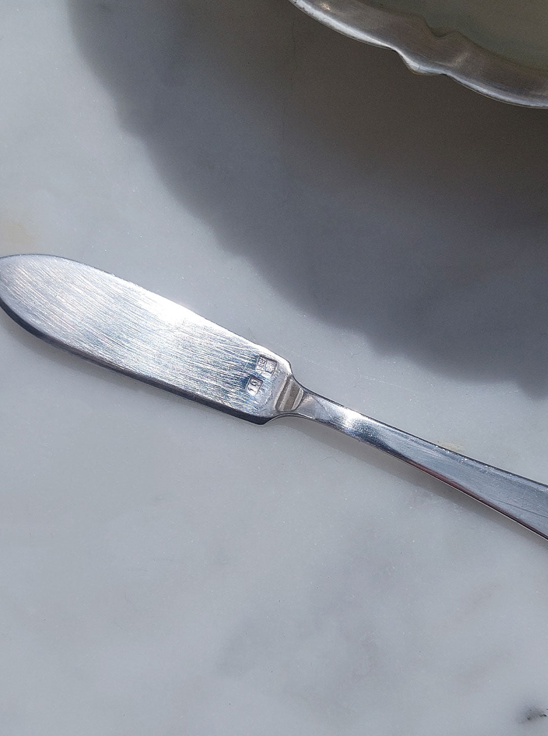 A close-up image of a butter knife resting on a light-colored surface. The metal knife, resting beside a Les Objoies Shell Silver-Plated Butter Dish with a seashell shape, has a smooth, reflective blade with a slightly rounded tip and a handle that tapers towards the end. The knife's surface displays some light reflections.