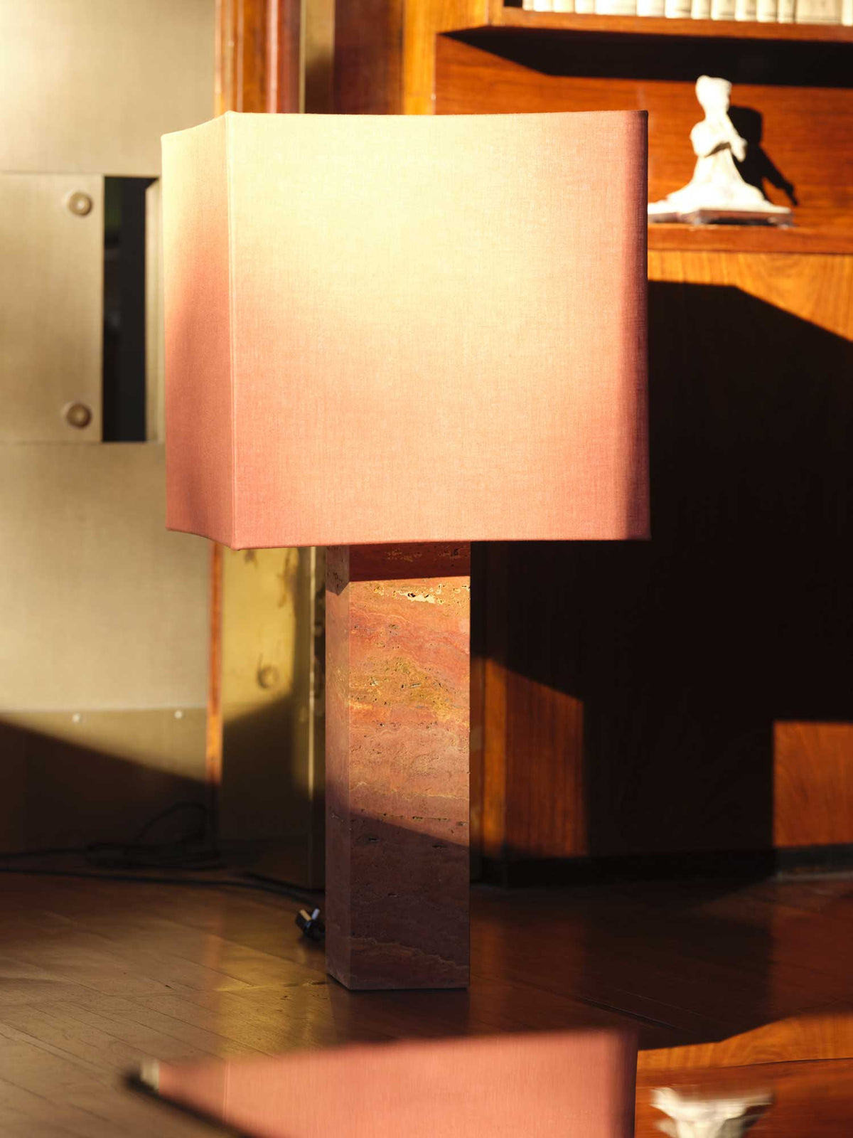 A stylish table lamp from Nassi Lamps, the Roma I Squared Red Travertine, is positioned in a room with wooden furniture and a bookshelf in the background. Natural materials blend seamlessly as sunlight casts a warm glow on the lamp and nearby surfaces, embodying elements of modernism.