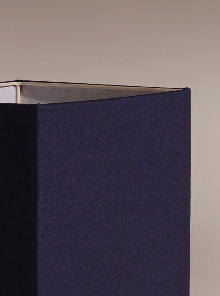 A close-up photo of a dark navy box slightly open at the top, revealing an off-white interior reminiscent of Nassi Lamps' Roma I Squared White Travertine. The background is a plain taupe color.