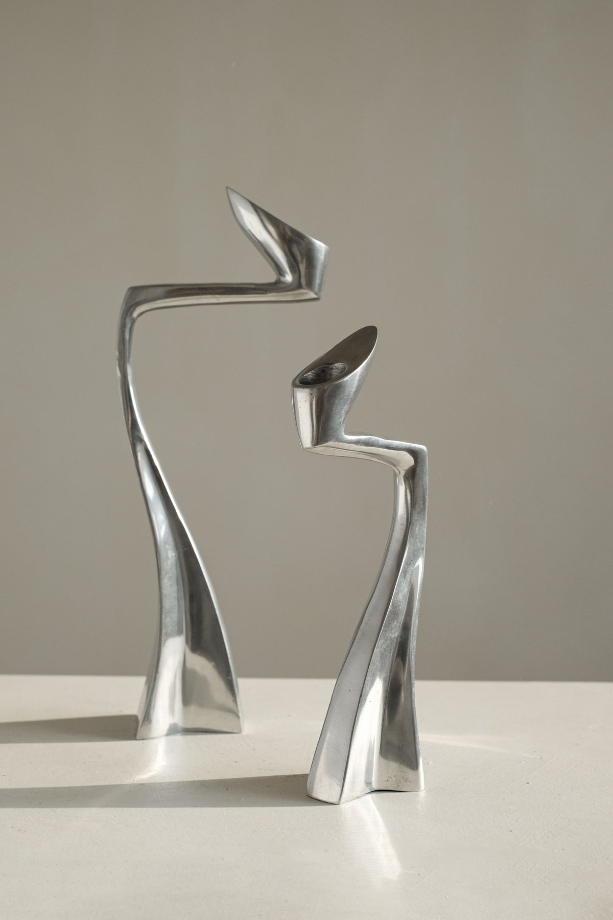 Two sleek, abstract candleholders from septembre studios stand on a flat surface. These collector’s pieces, with their elongated bodies and twisted shapes, evoke a sense of movement and balance against a plain background, captivating design enthusiasts.