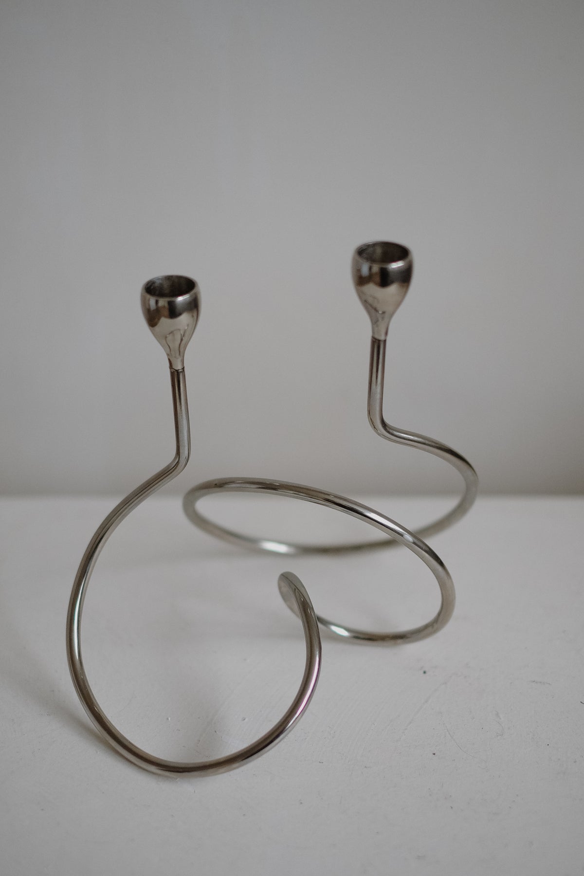 The "Loop" Candle Holder by septembre studios is a minimalist, spiral-shaped chrome design with two arms ending in candle cups. It rests on a white surface against a plain background.