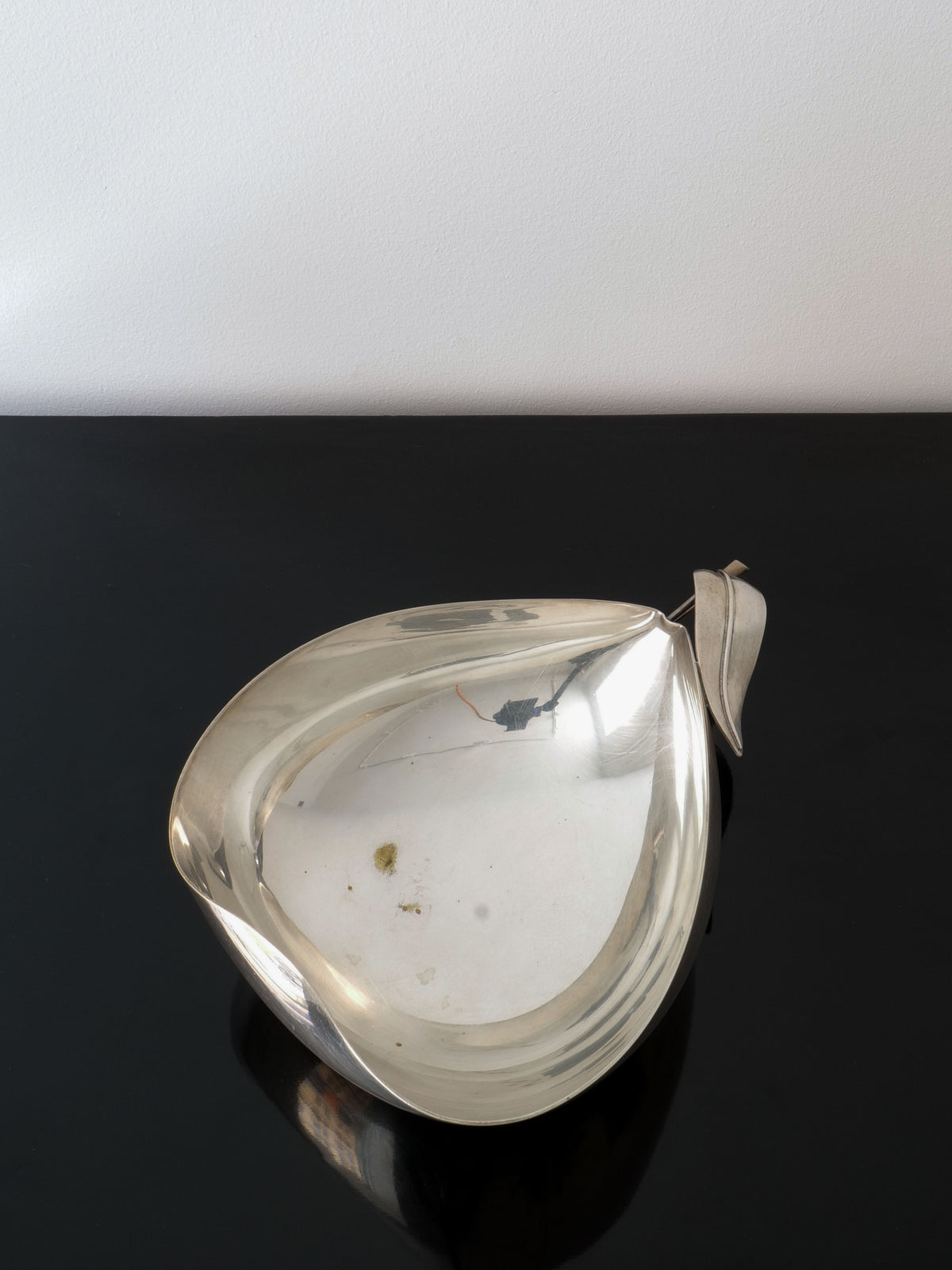 The Christofle Vide Poche Lino Sabattini by Collection apart is a silver, pear-shaped dish with smooth, shiny surfaces that sits on a reflective black table. This exquisite piece, designed by the renowned Lino Sabattini, features a small leaf detail at the top, resembling a pear stem.
