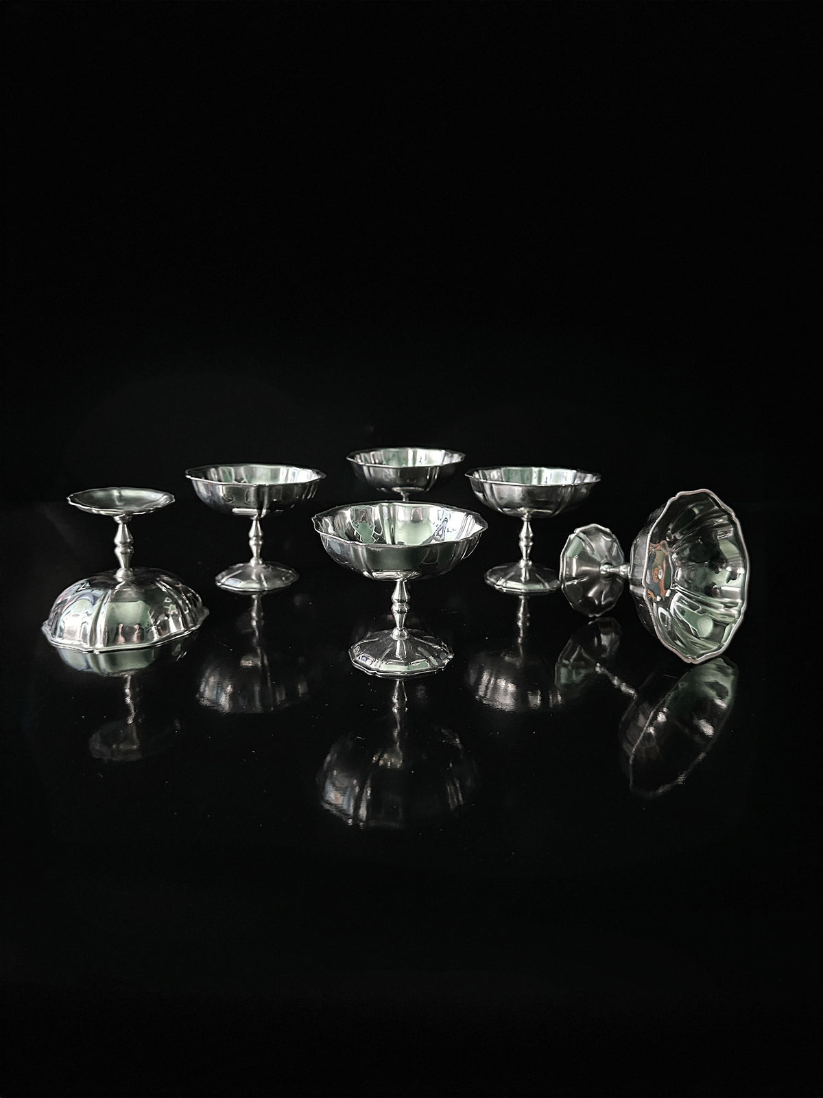 A set of seven Italian Stainless Steel Dessert Coupes by Dodo Vintage is displayed on a glossy black surface. Each dish has a reflective finish, creating mirrored reflections against a dark backdrop, highlighting the shiny texture of these elegant pieces.