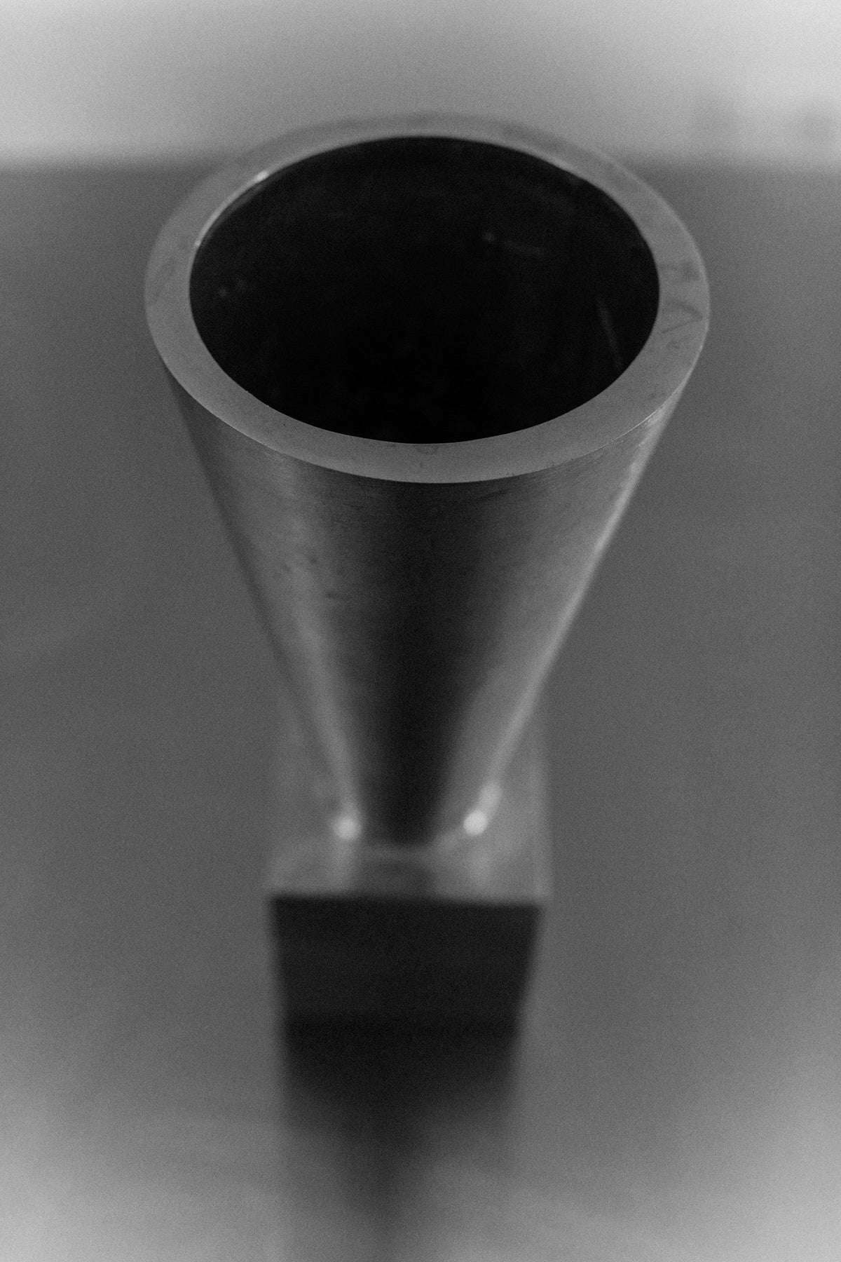 A black and white image features the 70's Aluminum Cast Vase by Out For Lunch. This abstract metal sculpture, crafted from brushed aluminum, tapers elegantly as it rises, resembling a minimalist vase. It is mounted on a rectangular base and positioned on a reflective surface that enhances its modern design.