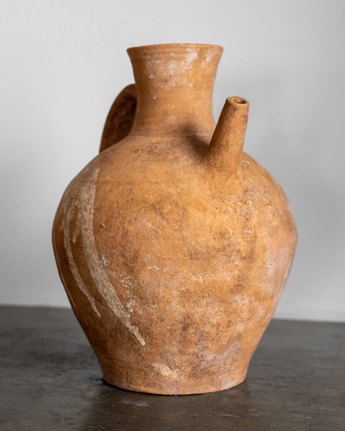 Spigel's Early 20th Century European Ceramic Pitcher radiates the charm of handcrafted pottery. It features a rustic brown color, wide body, narrow neck, one handle, and side spout with a rough texture. The pitcher is displayed gracefully on a gray surface against a plain white background.