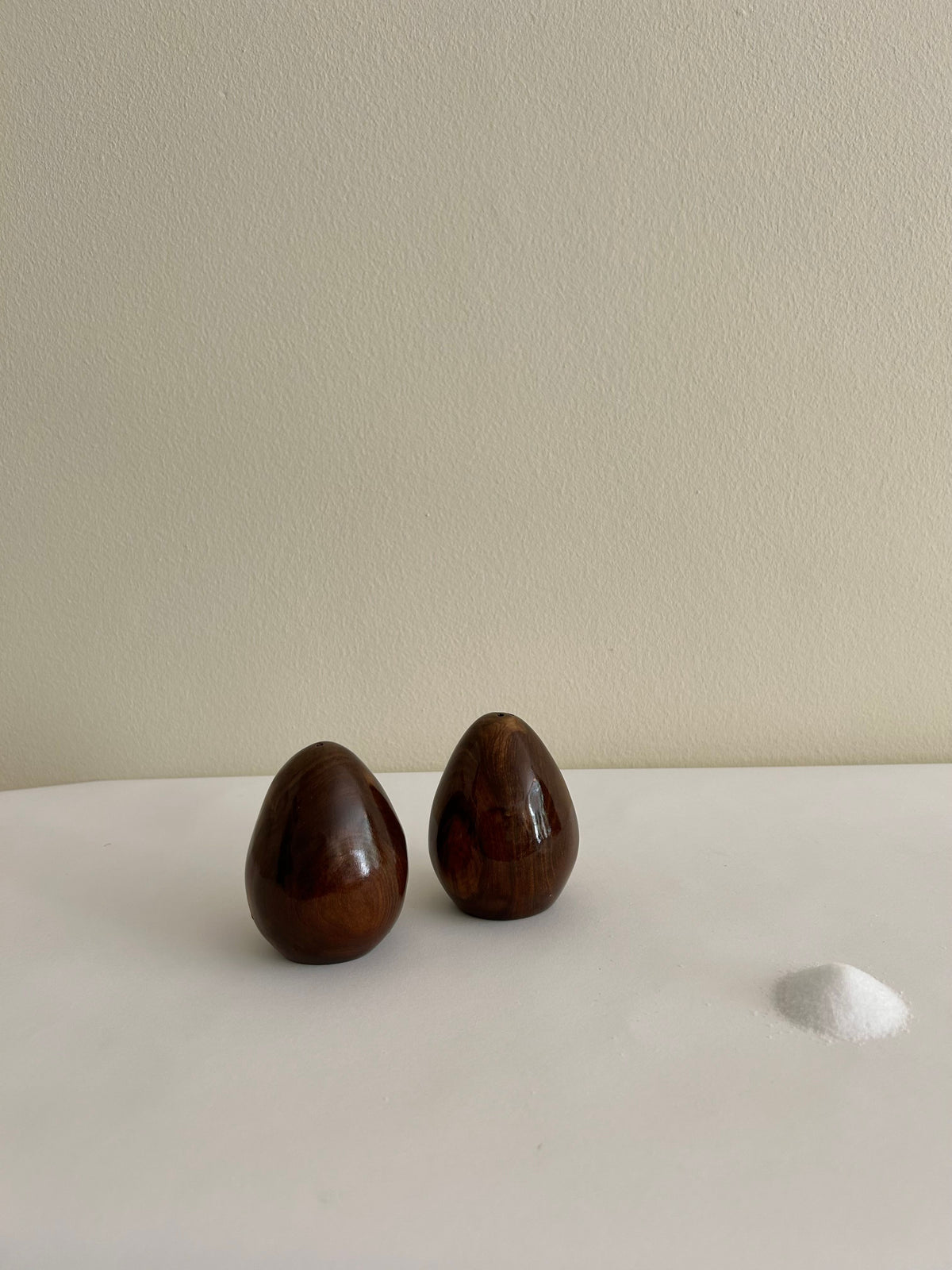 The Vintage Danish Wooden Salt & Pepper Set from Flock Studio, featuring two polished dark brown wooden eggs, is displayed like treasures in a vintage tableware collection on a white surface next to a small pile of white sand, set against a neutral light-colored wall.