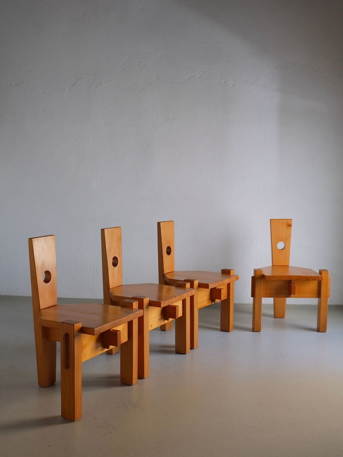 Four 1960s Erwin Egel-inspired pine chairs from Veter Vintage are arranged in a spacious room with a smooth gray wall. Each chair has a rectangular backrest featuring a circular cut-out near the top, and sunlight casts soft shadows on the floor.