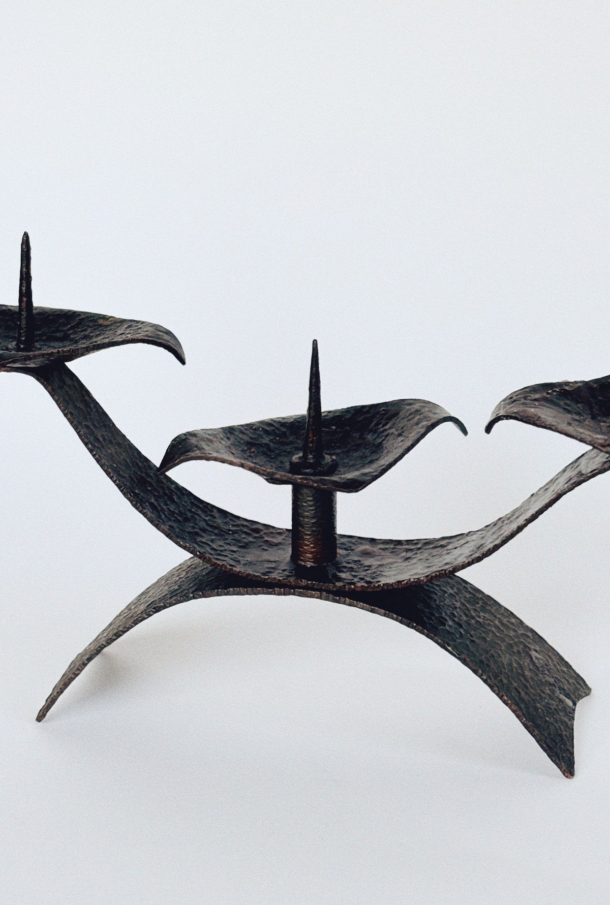 The Huerto 3 Arm Metal Candleholder features an ornate sculptural design with three curved platforms, each equipped with a central spike. This candlestick boasts artisan-made mid-century craftsmanship in a textured dark metallic finish.