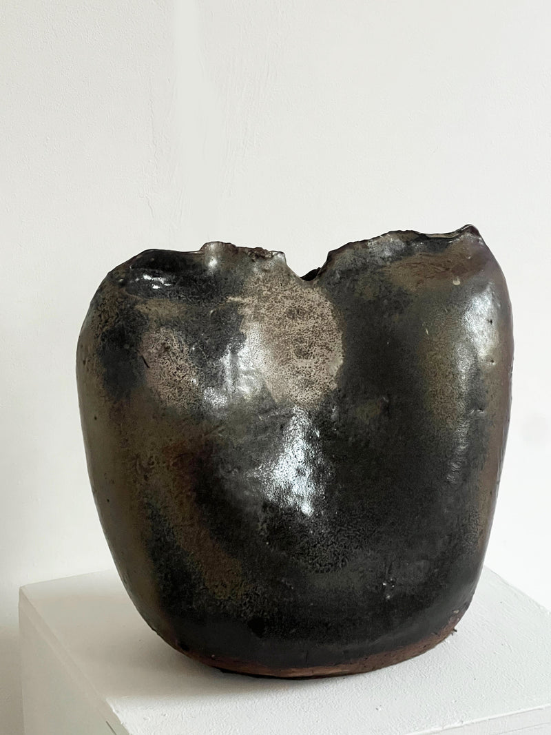 The Ceramic Pinch Top Studio Vase by Porter Studio, with its dark, textured finish and organic form featuring a roughly indented rim, is displayed on a white pedestal against a plain light background.