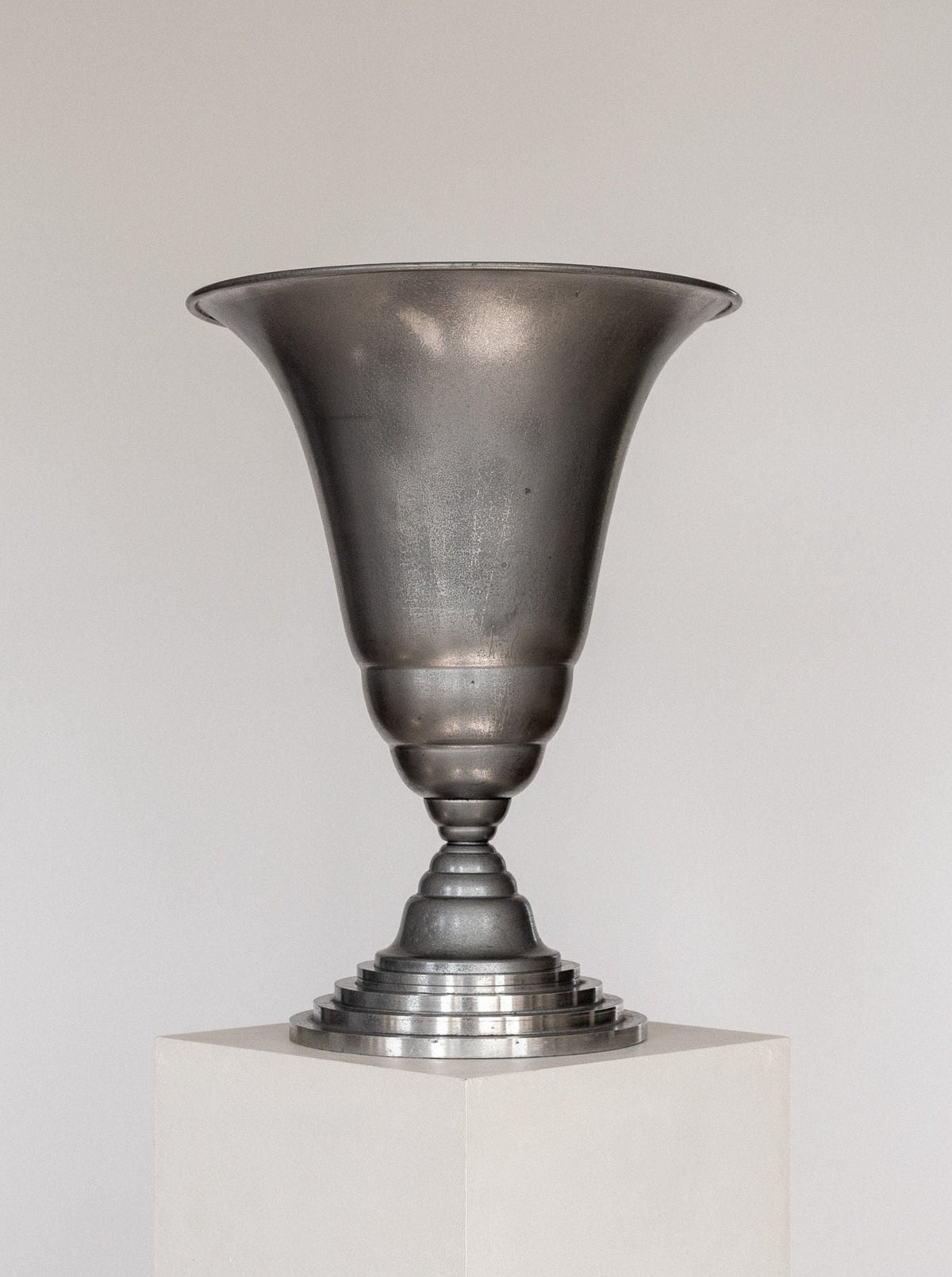 The Art Deco Torchière Table Lamp by Spigel, featuring a wide, flared brim, a narrow base, and a tiered stand, sits atop a white pedestal against a plain background. The French-designed lamp has a slightly tarnished, brushed finish that exudes vintage elegance.