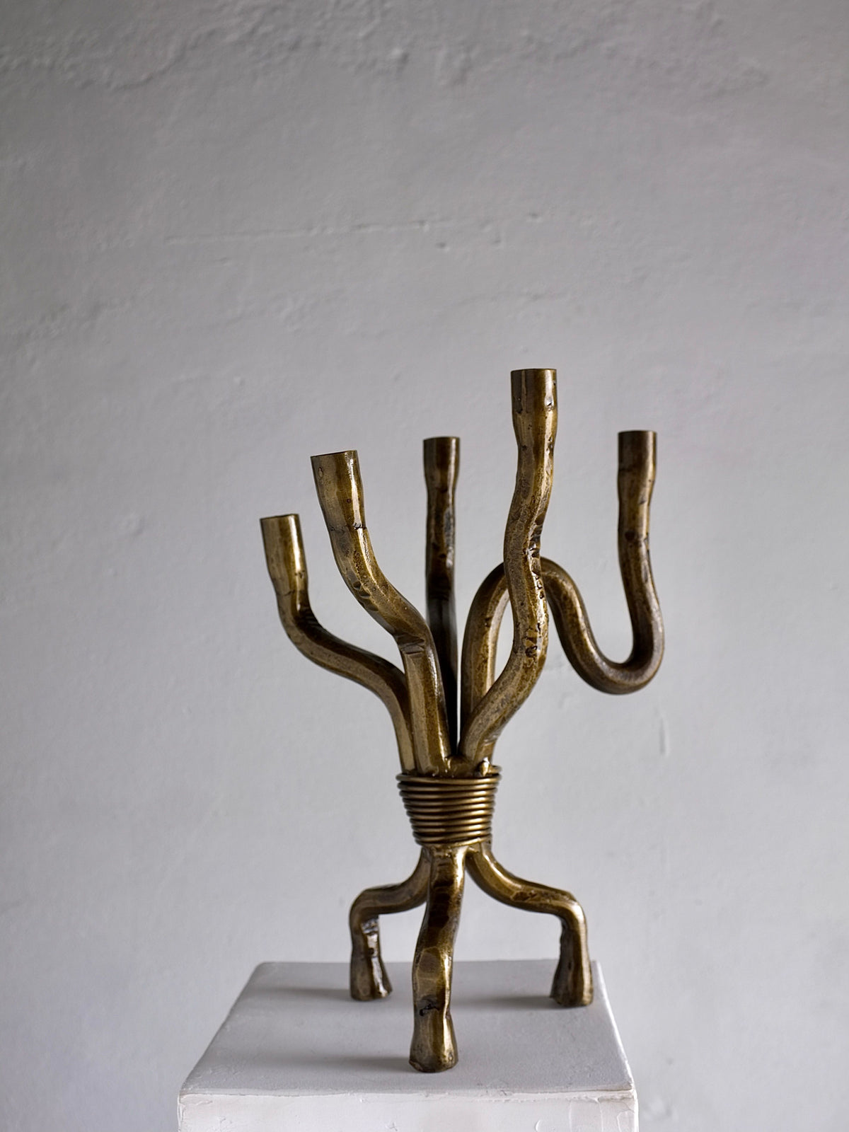 The Brutalist Brass Candelabra by Veter Vintage, crafted in the Netherlands during the 1970s, features a vintage design with six unevenly curved arms intended to hold candles. The sturdy base is supported by four legs and is showcased on a simple white pedestal against a plain gray wall.