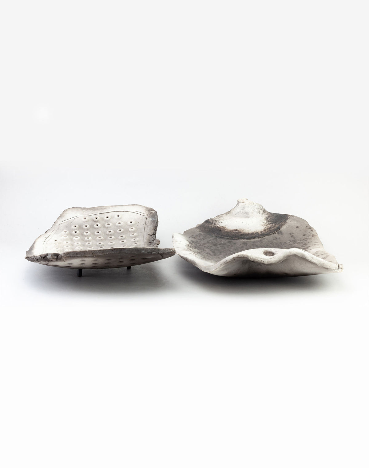 Displayed against a plain background are two freeform raku platters by John Dunn from RELIC LONDON. One sculpture is distinguished by its circular holes, while the other has a smooth design with wavy edges. Both pieces are primarily white with subtle gray accents, capturing a minimalist elegance.